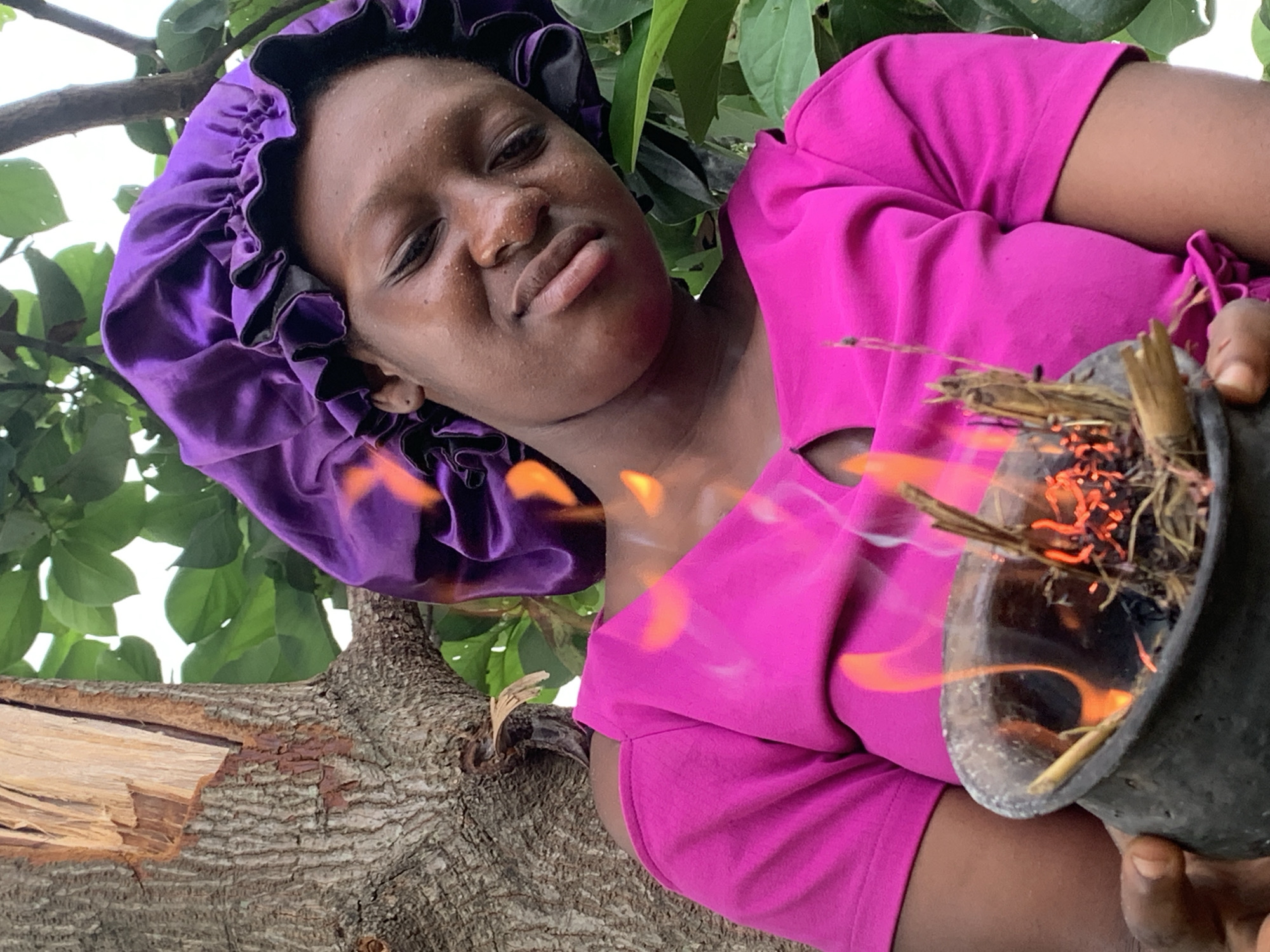 The image depicts a person with dark skin wearing a purple satin bonnet and a magenta top, holding a burning pot. The pot contains sticks and embers, with visible flames emanating from it. The person is outdoors, standing near a tree with green foliage and a broken branch. The location is claimed to be Agban, Nigeria, though there are no visual cues to confirm this.