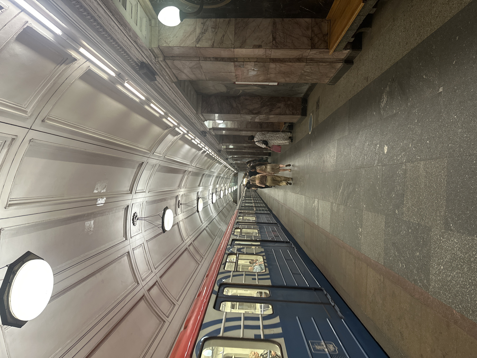 This image shows a Moscow Metro station. The station features a high, arched ceiling with intricate white paneling and a series of decorative lights. Along the platform, a blue and red train is stopped, with several passengers visible through the windows. The platform itself is paved with dark stone and a lighter brown border runs along the edge near the train. On the right side of the platform, there are marble-clad pillars and a wooden bench. Several people are walking along the platform. The lighting suggests it is daytime.