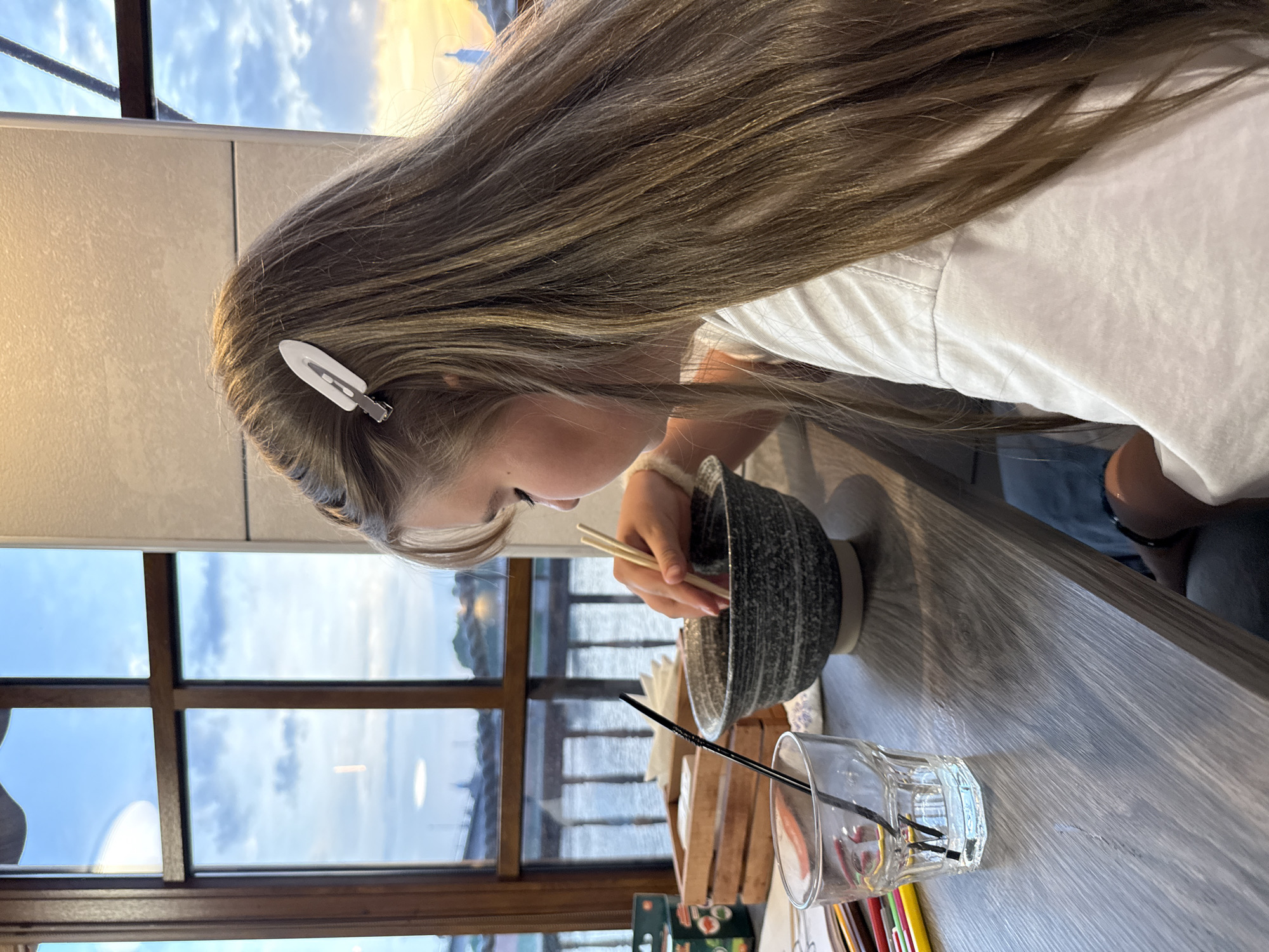 A fair-skinned, young, light-haired girl sits at a table next to a window, facing left, eating from a gray bowl with chopsticks. A white hair clip is in her hair, and she is wearing a white top. In front of her is a clear glass with a black straw and several pencils. Through the window, a body of water and buildings are visible. Based on the architecture and the time of day, the location is potentially the city of Veliky Novgorod, Russia, near the Volkhov River.