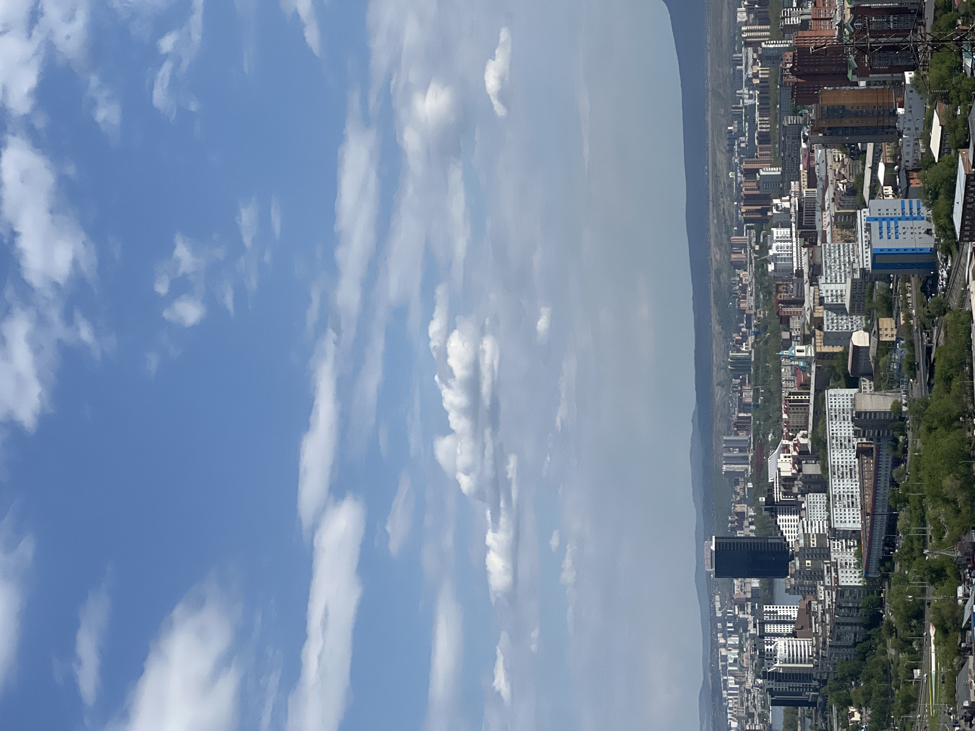 The image shows a cityscape in Krasnoyarsk, Russia, beneath a blue sky with scattered clouds. The city is dense with buildings of varying heights and architectural styles, ranging from modern high-rises to older, smaller structures. A river runs through the city. In the background, a mountain range is visible, veiled by atmospheric haze. The sky is mostly clear with thin, elongated clouds.