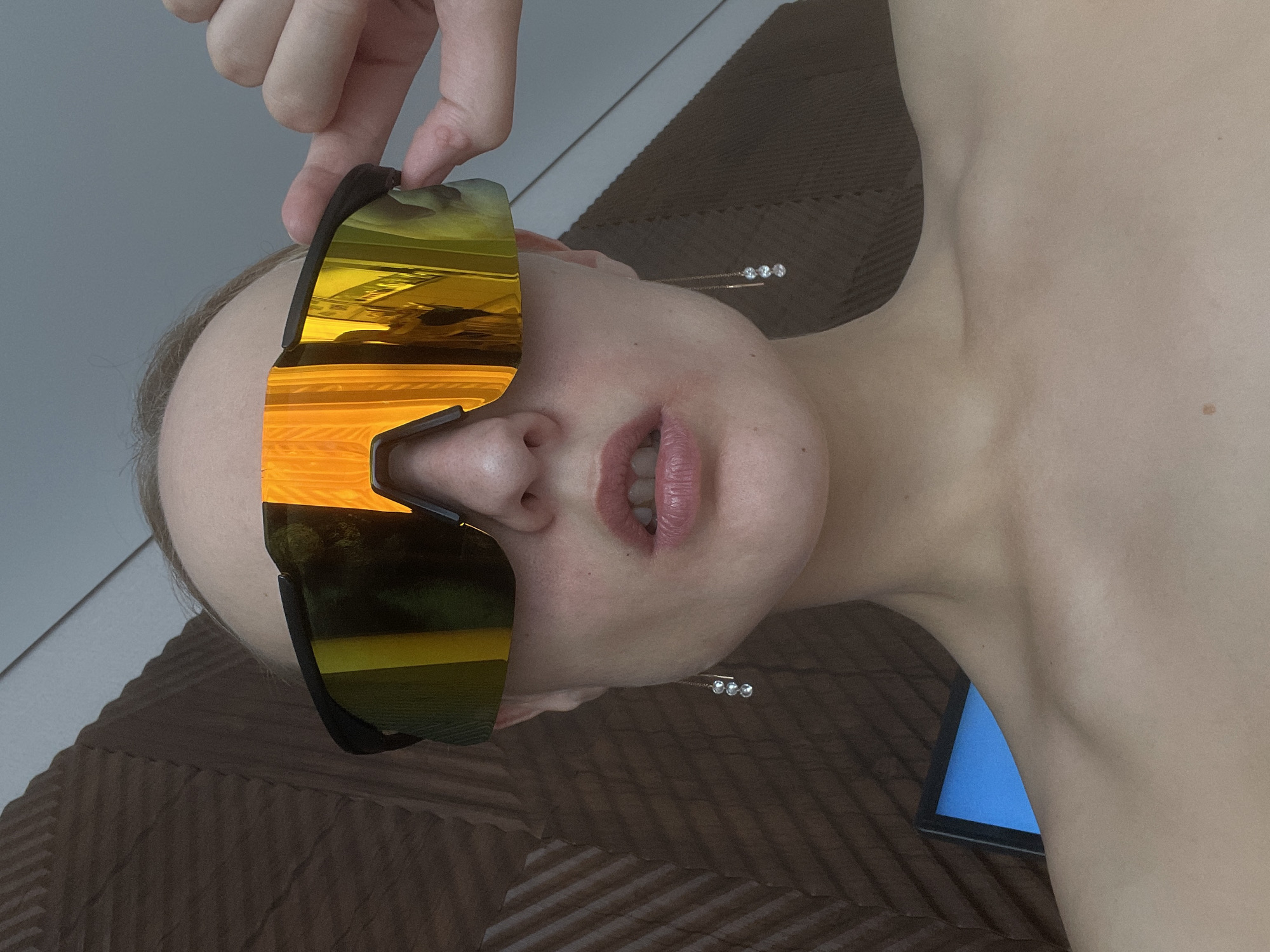 The image shows a person lying on a brown textured surface, likely a bed or lounge chair, with part of a tablet visible near the neck. The person is wearing large, reflective sunglasses with a black frame, held by a hand adjusting them at the top. The reflection in the sunglasses shows an outdoor scene with green vegetation and a golden or orange light, potentially a sunset. The person's face, including the lips, nose, and one ear with a dangling earring, is visible beneath the sunglasses. The image does not contain enough information to confirm the location is Подшивалово, Russia. The scene is generally still.