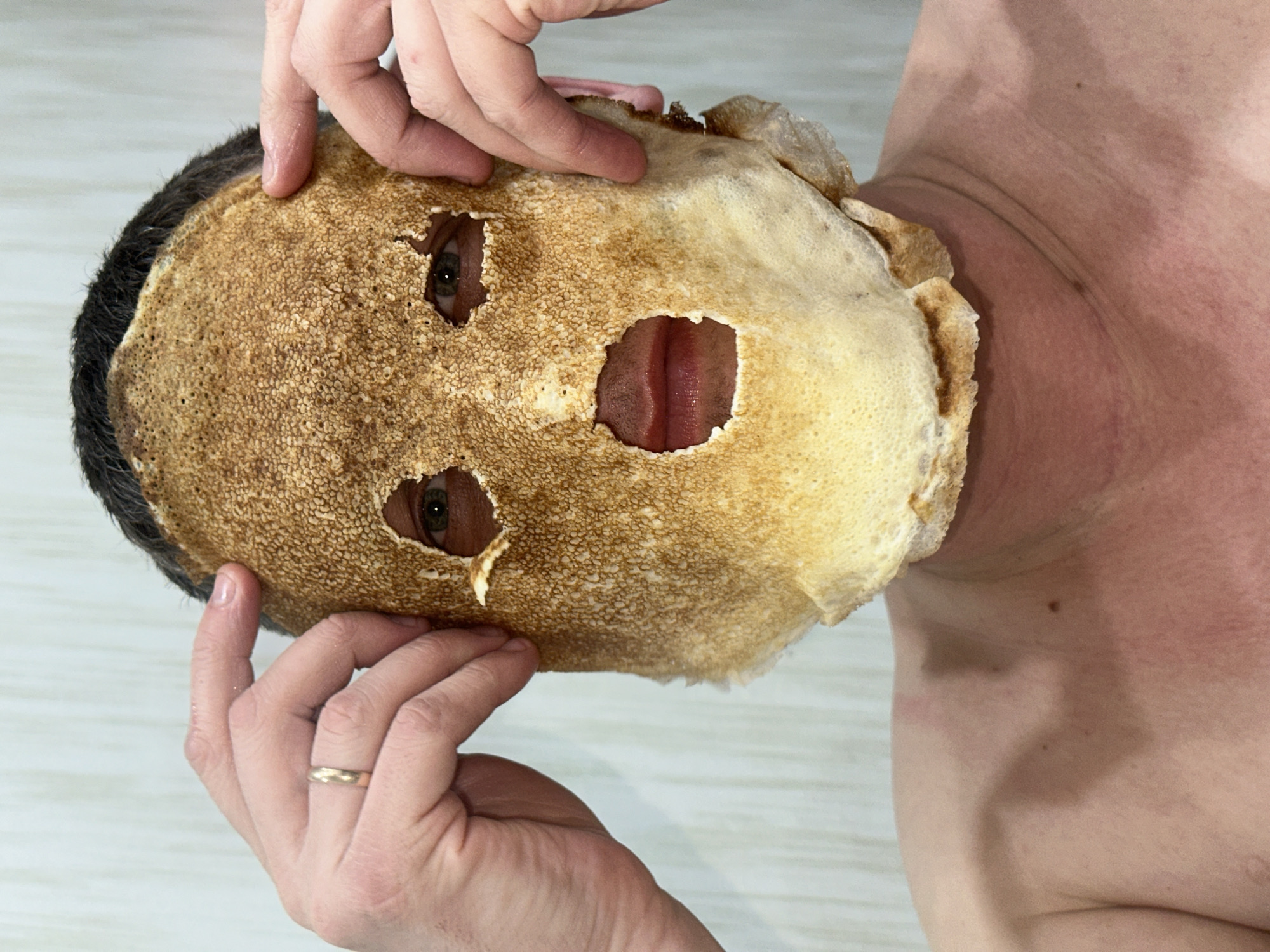 A man's face is partially covered by what appears to be a browned crepe with holes cut out for the eyes and mouth. His head is tilted to the right. Two hands are positioned on either side of the crepe, holding it in place. The man has dark hair and visible skin on his upper torso. A wedding band is visible on the ring finger of one of the hands. The background is indistinct, appearing to be a plain white or light-colored surface. There is no visual evidence that specifies that the location is in the city of Chaikovsky, Russia.