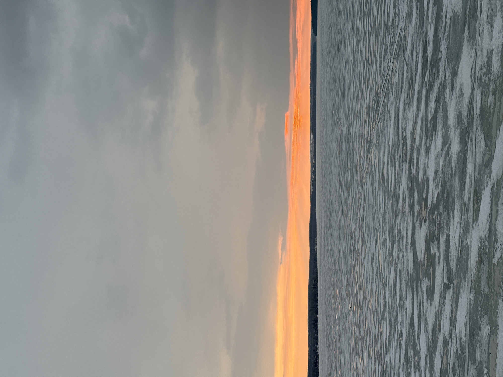 The image depicts a landscape at the location of the city of Votkinsk, Russia, during a sunset over a body of water. The sky is predominantly covered with overcast grey clouds, contrasting with a vibrant orange and yellow glow on the horizon, likely due to the setting sun. The land or far bank of the body of water is a dark, almost black line at the base of the horizon. The water itself exhibits a textured surface, possibly indicating ice or a combination of ice and water. The texture creates a pattern of light and dark areas, contributing to the overall contrast in the image. There are no visible people or animals, focusing solely on the natural scene.