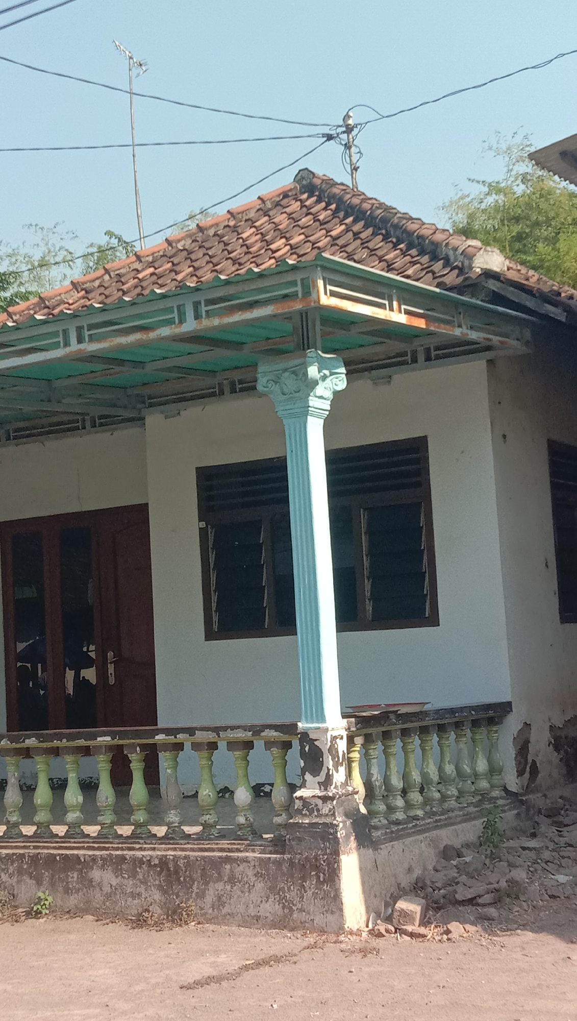 The image shows a weathered house with a tiled roof and a covered porch supported by a light blue pillar. The house has white walls and dark brown double doors. There are horizontal sliding windows on the house's exterior. A green and white ornate railing runs along the porch, with some sections showing paint peeling and damage. The ground in front of the house is paved and dusty, with some debris and small plants visible. In the background, there are trees and a clear blue sky. An antenna is visible on the roof, and electrical wires run across the sky. The house appears to be located in Sidogiri, Indonesia.