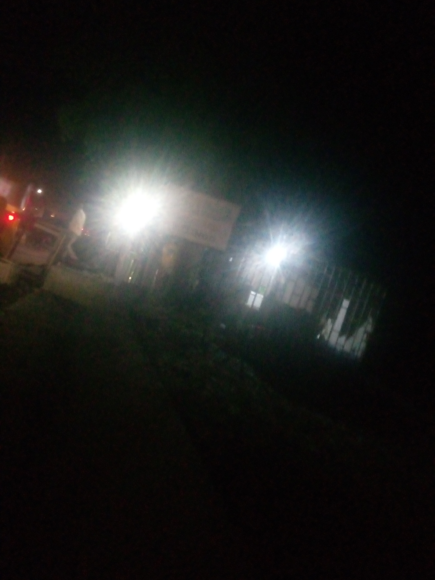 This image appears to be taken at night, possibly in Jalingo, Nigeria. The scene is dimly lit, with artificial lights creating bright flares and reflections. In the foreground, there is dark foliage or vegetation. In the mid-ground, a structure with what appear to be barred windows or a fence is visible, with some interior lights illuminating the space. There are also vehicles present, indicated by headlights and red taillights. One particularly bright light source, possibly a street lamp or floodlight, is prominently featured. The overall impression is that of a street or a building exterior at night, with limited visibility and significant light distortion.