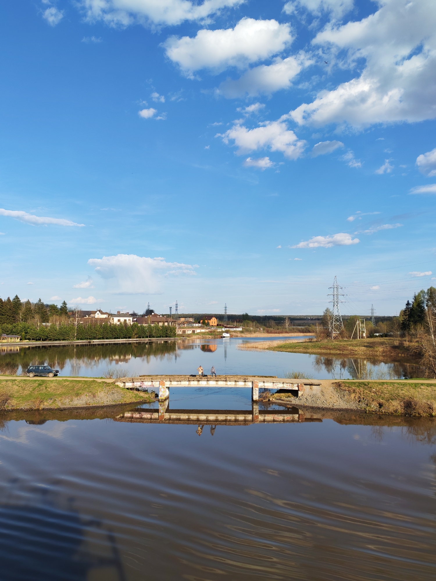 The image depicts a rural scene under a bright blue sky with scattered white clouds. The scene is dominated by a body of water, likely a river or a wide canal, with its surface showing slight ripples. A small concrete bridge spans the water, connecting two strips of land on either side. Two individuals are visible on the bridge.
On the left side of the bridge, a dark-colored vehicle, possibly a car, is parked on the grassy bank. Beyond the water and bridge, there is a row of buildings, possibly houses or small industrial structures, with varying rooflines and colors. Utility poles with power lines are also visible in the background. The landscape includes trees and vegetation.