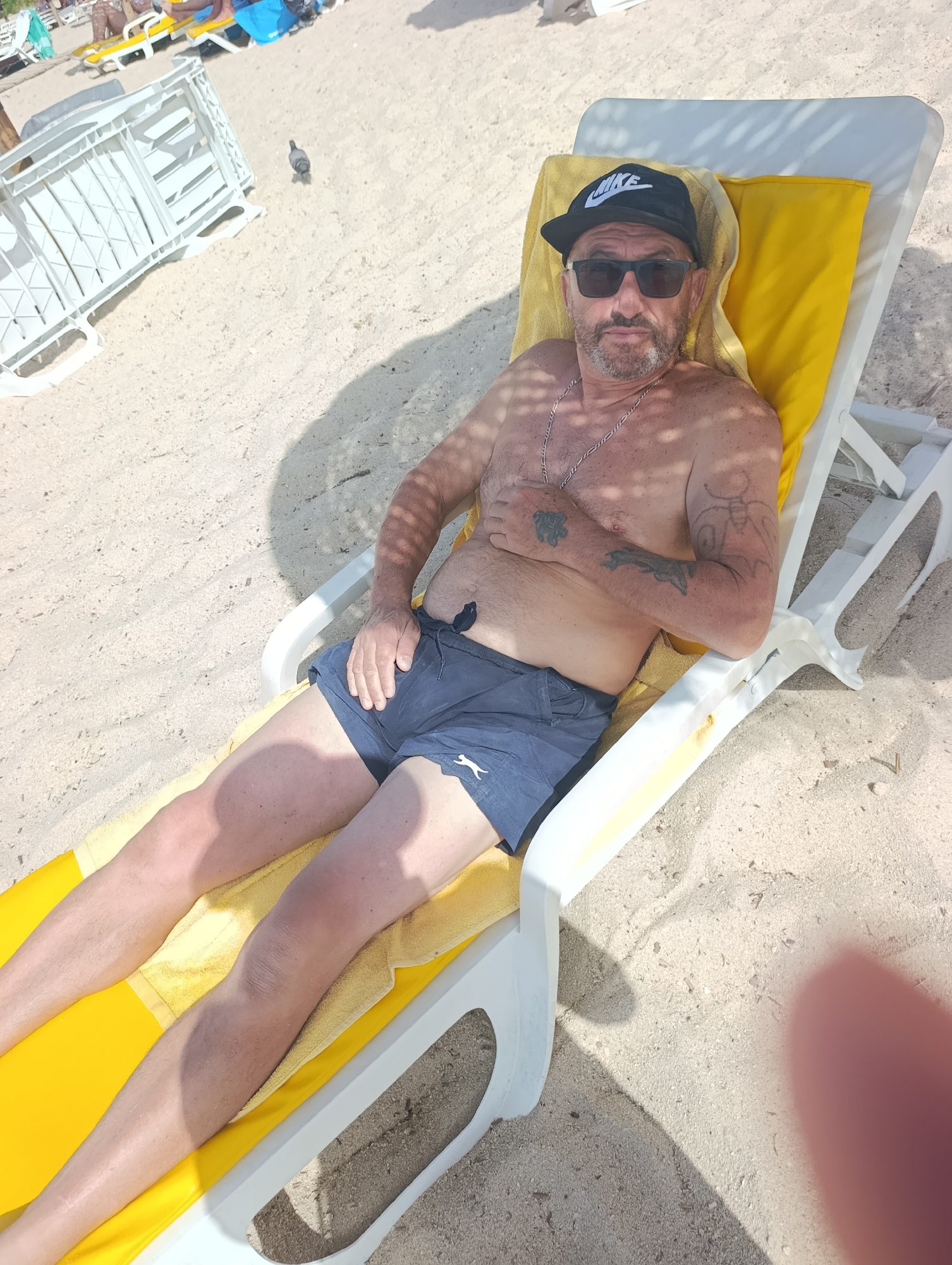 A man wearing a black baseball cap and sunglasses lies on a yellow towel on a white lounge chair on a sandy beach. He is shirtless, wearing dark blue swim shorts. The man has tattoos on his arms. The beach is lined with lounge chairs and umbrellas, some occupied by people in the background. The man is looking at the camera. The scene appears to be in a tourist area.