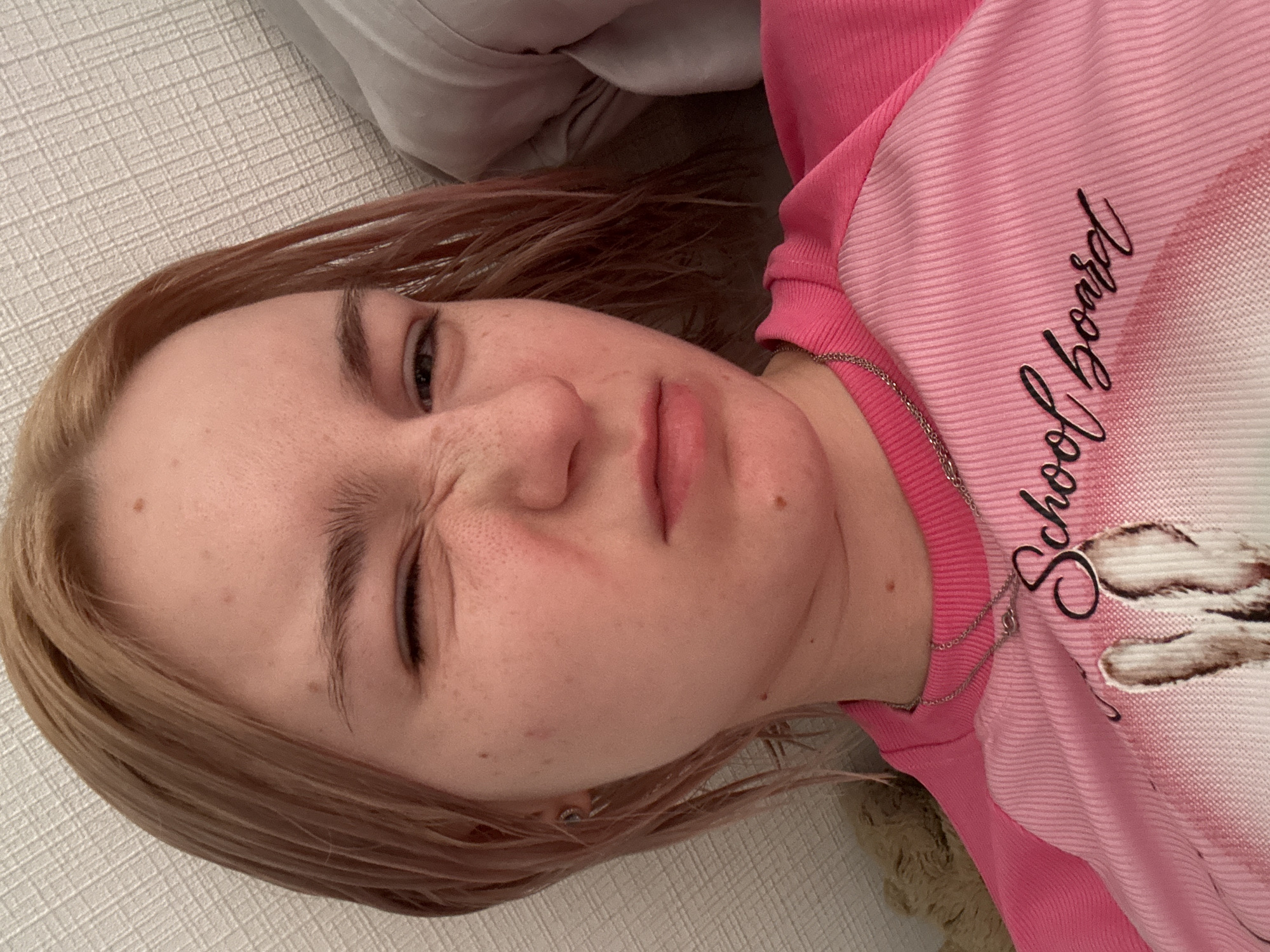 A close-up, high-angle portrait captures a young individual with an expressive facial contortion, appearing to be a selfie. The subject has light reddish-blonde hair, parted on the left side, with visible strands. Their skin is fair with numerous freckles across the forehead, nose, and cheeks, along with several small moles, including one prominently above the right eyebrow and another near the left corner of the mouth. The right eye (viewer's left) is tightly closed, forming deep wrinkles at the corner, while the left eye (viewer's right) is partially open and squinting. The nose is significantly scrunched, and the lips are slightly pursed and downturned. A small, silver-colored, abstractly shaped earring is visible in the left earlobe.
The individual wears a two-toned, long-sleeved shirt. The sleeves and collar are solid fuchsia pink with a fine ribbed texture. The body of the shirt is a lighter, pale pink, also ribbed, featuring a graphic print of light-colored, fluffy animal ears (possibly a rabbit) at the bottom of the frame. Overlaid on this graphic, in a black script font, are the words "School board." A thin, silver-colored chain necklace with a small, indistinct pendant is worn around the neck.
The background consists of a light-colored, textured wall (possibly wallpaper with a grid-like pattern) on the left side, and soft, light grey fabric (likely a pillow or blanket) on the right side behind the individual. A small, dark brown, plush object is partially visible in the bottom left corner. The scene is located in the city of Unknown, Russia.