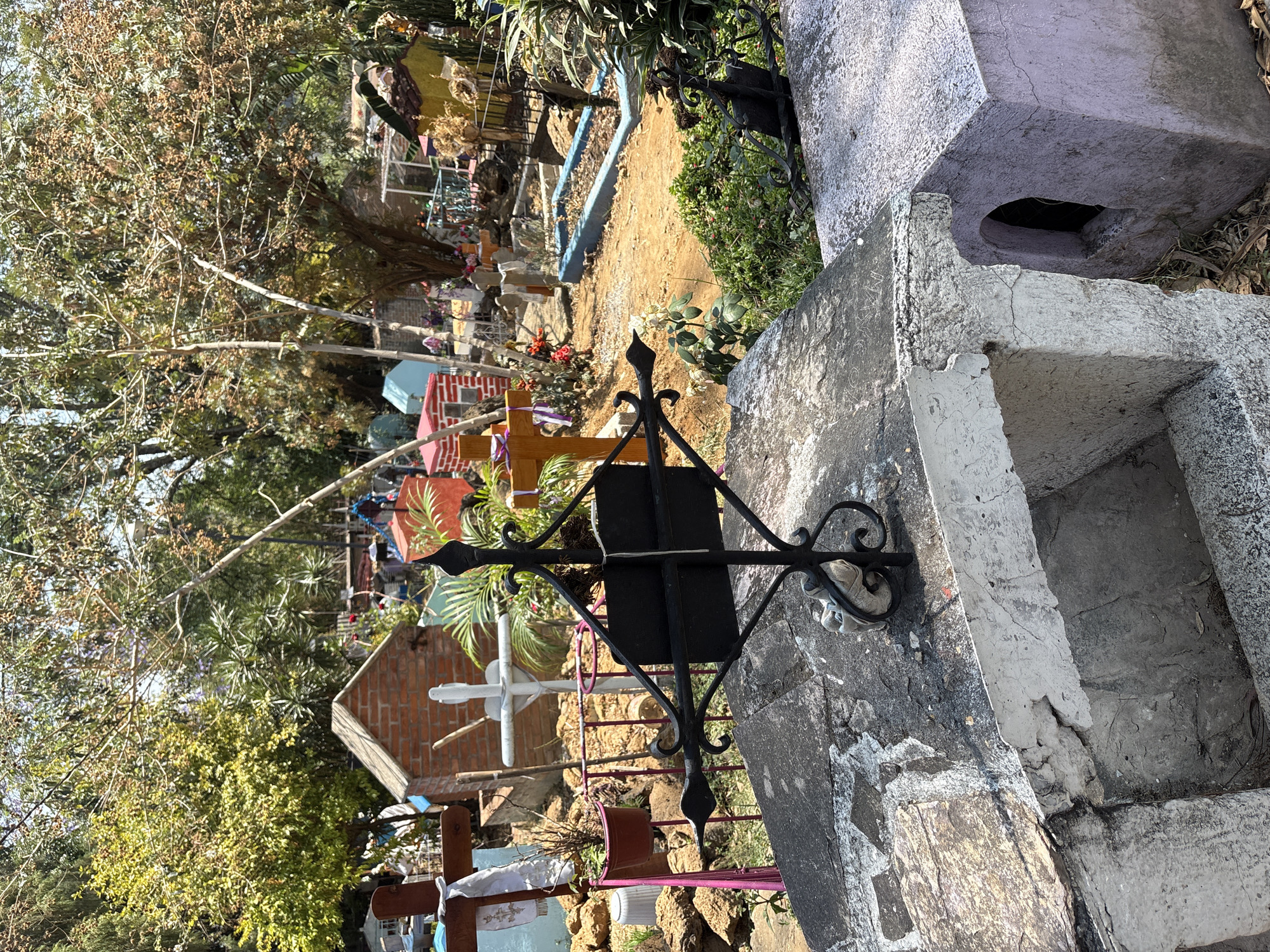 The image shows a section of a cemetery in Ocotepec, Mexico. In the foreground, there are grave structures made of concrete and stone, one adorned with a black wrought iron cross. The graves show signs of weathering and age. Beyond the immediate foreground, the cemetery extends, featuring a variety of graves marked with crosses, flowers, and colorful decorations. Vegetation, including trees and shrubs, is interspersed throughout the scene. The background suggests a vibrant, populated burial ground.