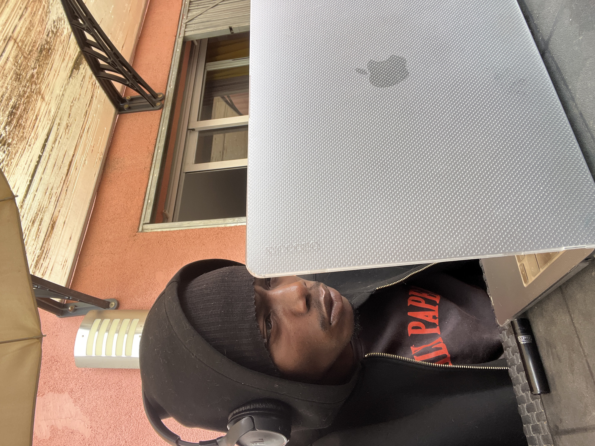 This rotated image captures a person sitting outdoors with a laptop, set against a building facade in Barcelona, Spain. The subject is a man with dark skin, wearing a black beanie and a black hooded sweatshirt, with black headphones resting around his neck. He has a neutral expression, looking towards the camera.
In front of him is an open laptop, likely a MacBook, covered by a clear, textured protective case that faintly displays the "MacBook" brand on its top left. The laptop screen is not visible. To the right of the laptop, a black Clipper lighter with "Clipper" and "reusable" text is visible on a dark, textured surface, possibly a table. Partially visible on the man's shirt, beneath his open hoodie, is some red text, appearing to read "A L PAP" or similar.
The background features a reddish-orange stucco building wall. A window with a white metal frame and partially closed shutters is prominent. Above the window, a dark metal bracket supports an awning or sunshade, part of which is visible on the left. A silver-toned outdoor light fixture with vertical light strips is mounted on the wall. The scene is illuminated by natural daylight, suggesting it is daytime.
