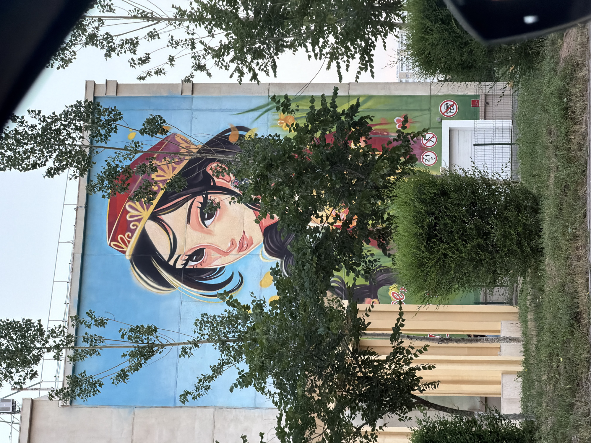 A mural of a young woman wearing a traditional red and gold hat adorns the side of a building in Astana, Kazakhstan. The woman's face is depicted in a stylized anime-like manner, with large expressive eyes and dark hair. She is surrounded by painted flowers and greenery against a bright blue sky. In the foreground, several trees and bushes partially obscure the mural. The building itself is painted in muted tones with yellow columns visible on the left side. A white garage door is visible at the base of the building, along with several signs. The overall scene suggests an outdoor urban environment.
