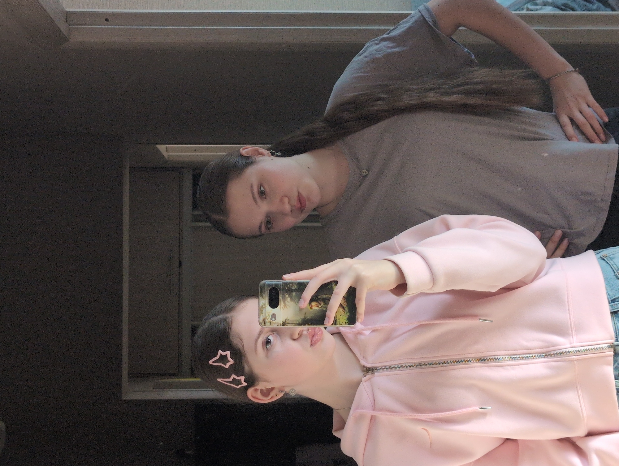 Two young women are visible in a mirrored selfie. One, wearing a pink zip-up hoodie and star-shaped hair clips, is holding a phone with a landscape scene printed on the case. She has light skin and light hair, and she is pursing her lips in a "duck face" expression. The other young woman is reflected in the background. She is wearing a gray t-shirt and has long brown hair. There is a light fixture and a closed cabinet door visible in the background of the reflection. Without more information, I cannot determine if this photo was taken in Izhevsk, Russia.