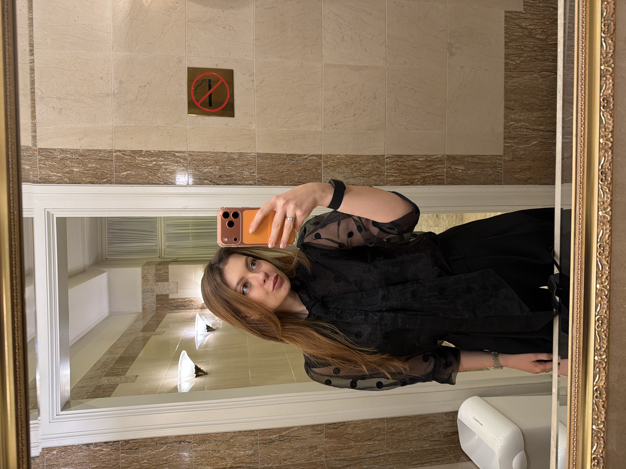 A woman is taking a mirror selfie in a bathroom. The background features beige tiled walls with a brown horizontal accent and a "No Smoking" sign. The mirror has an ornate gold frame. The room visible through the mirror seems to be a hallway. The woman is wearing a black polka-dot mesh blouse. A hand dryer is seen at the bottom right. This may be inside the Metropol Hotel in Moscow, Russia, due to the bathroom details.