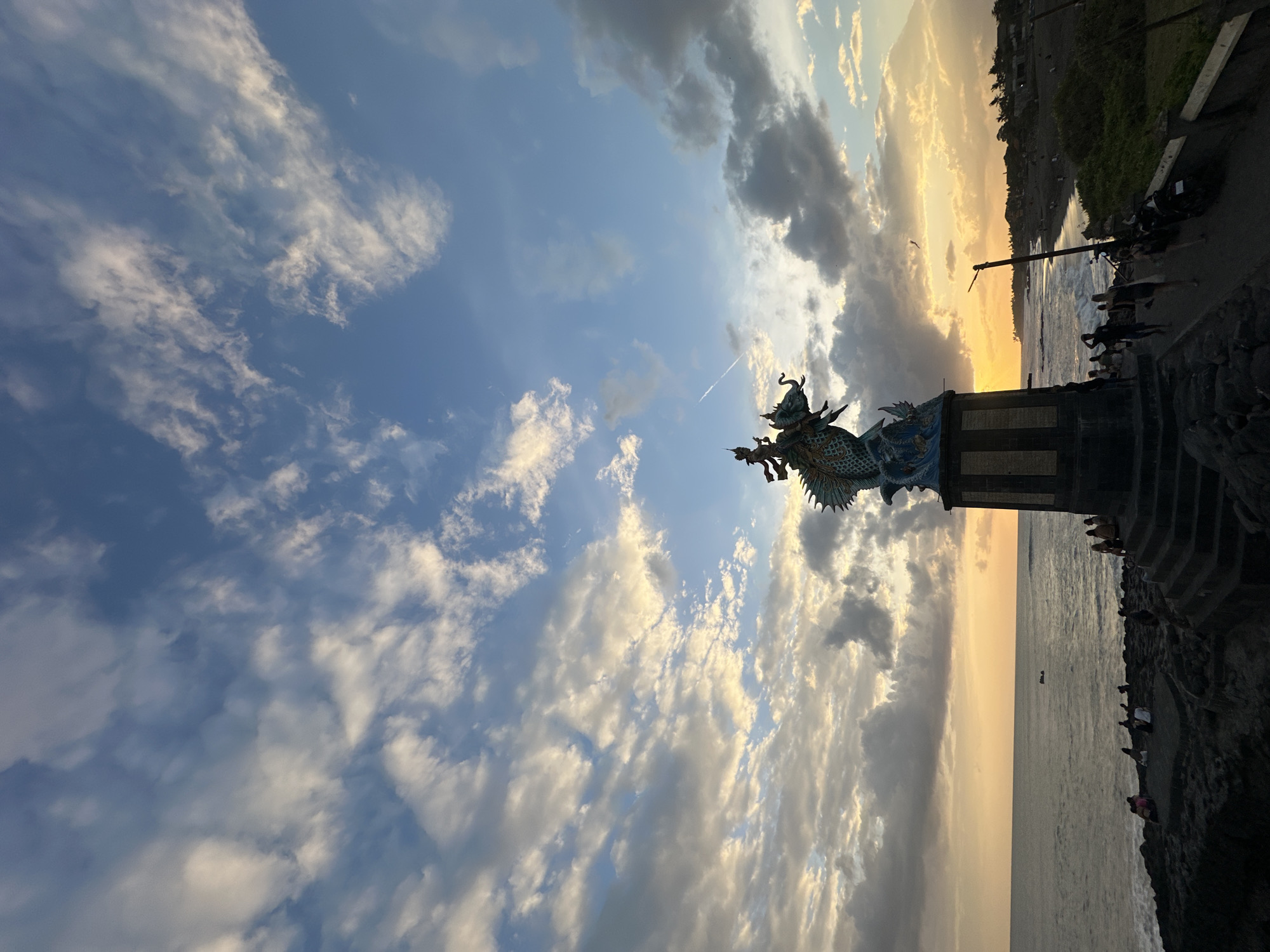 A statue of a mythical creature, possibly a dragon, adorned with a rider, stands prominently on a pedestal by the sea at sunset. The sky is a vibrant canvas of blue with scattered white and gray clouds, illuminated by the warm glow of the setting sun. The ocean stretches out to the horizon, with gentle waves lapping against the rocky shore. Several people are scattered along the coastline, some seated on the rocks, others walking, and a few are near the statue. A dark, sandy beach is visible in the background, lined with palm trees and buildings. The overall scene depicts a serene and picturesque coastal landscape with a touch of cultural or mythical symbolism.