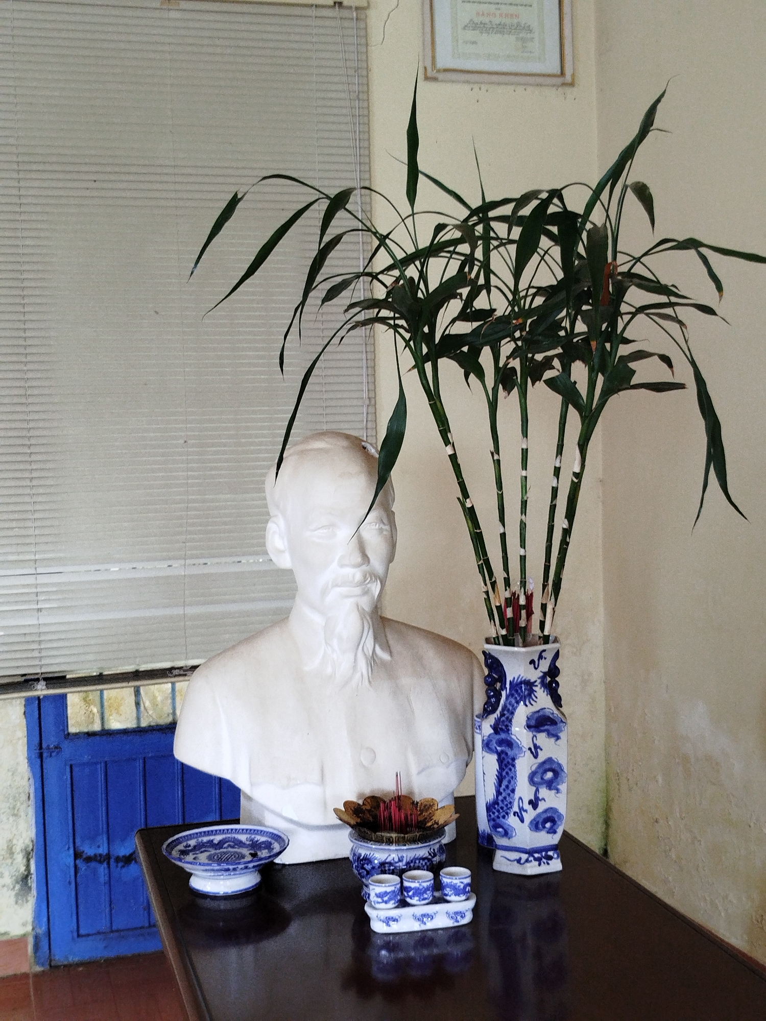 The image shows a bust of Ho Chi Minh, an assortment of incense holders and saucers, and a vase of bamboo stalks placed on a dark wood table. The room features a window with closed blinds, a framed certificate hanging on the wall, and a blue wooden door. The location is likely in Lang Biang - Đà Lạt, Vietnam.
