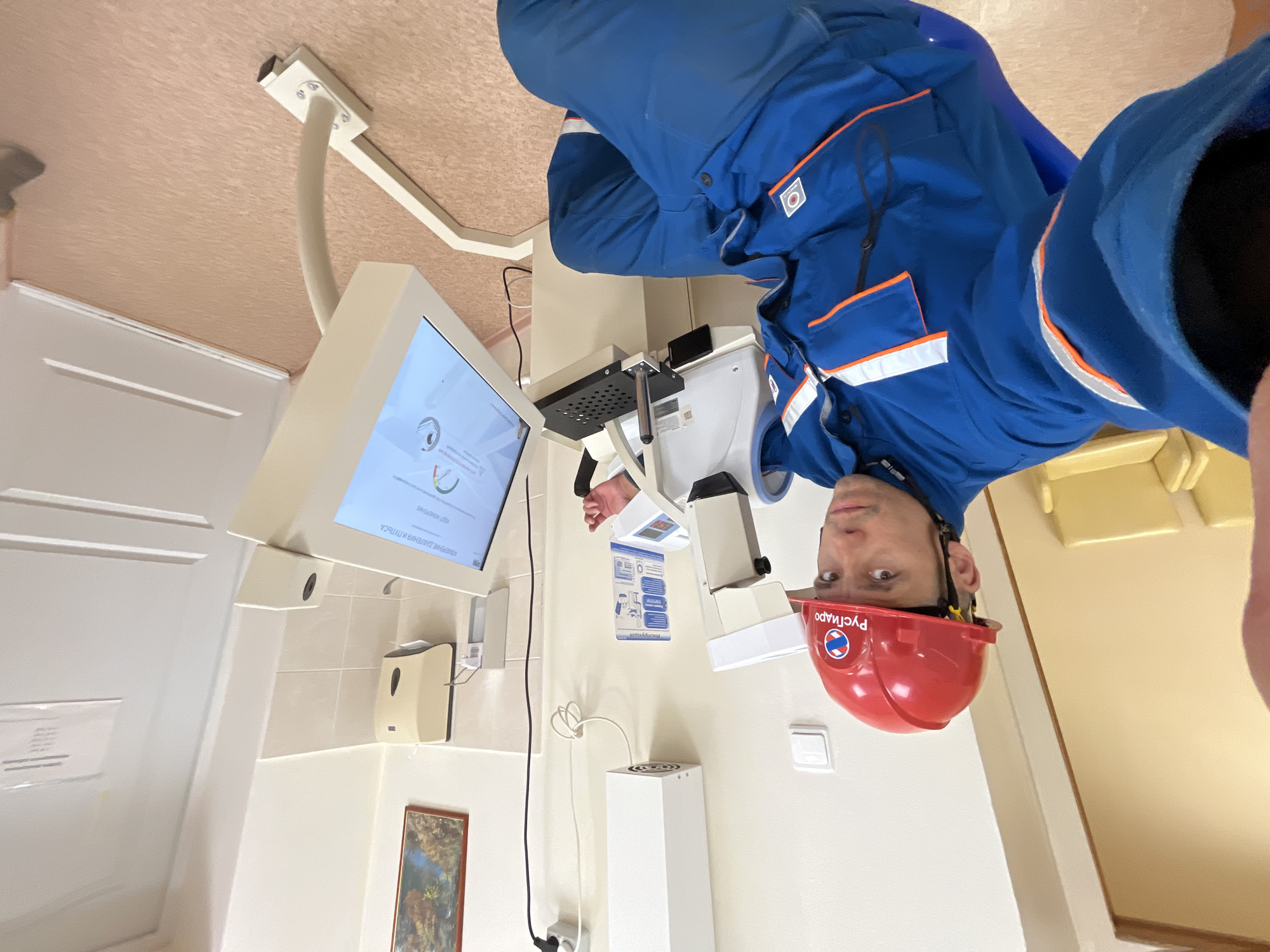 A man in a blue work suit and a red hard hat with "Русгидро" (RusHydro) printed on it is in an examination room, possibly a medical clinic. He appears to be taking a selfie or recording a video. He is standing near a medical device with a screen showing text in Russian, which likely measures blood pressure and pulse. The examination room has white walls, a dispenser for hand sanitizer, a framed picture on the wall, and other medical equipment. The location could be a medical facility or a health check station in Chaikovsky, Russia, considering the writing on the hard hat and the language on the screen.