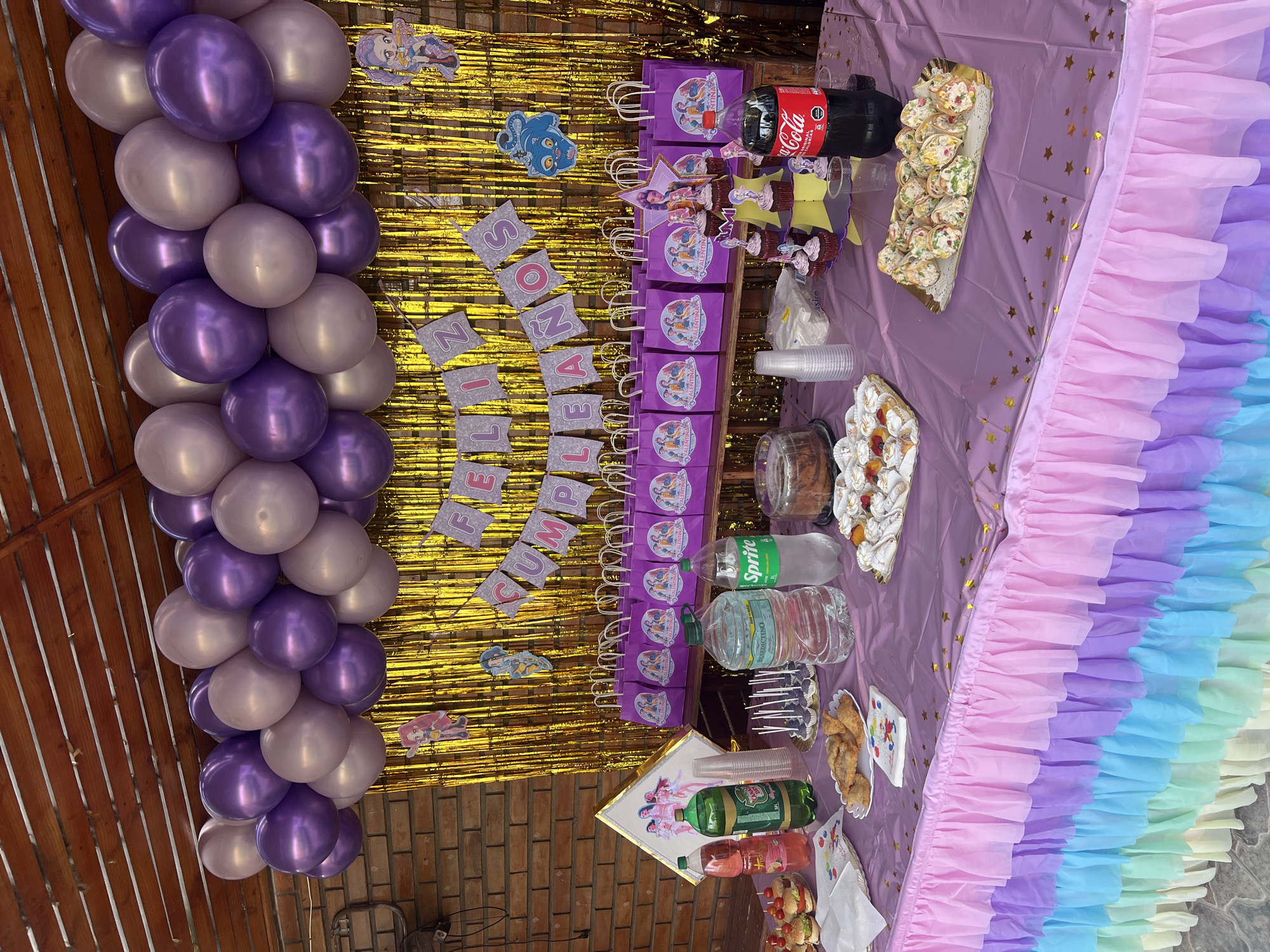A party scene is set outdoors in Arica, Chile, indicated by the brick wall. The party is decorated with a balloon garland of purple and silver balloons. A gold tinsel backdrop hangs behind a banner that reads "FELIZ CUMPLEAÑOS" which translates to "Happy Birthday." A table is covered with a purple tablecloth adorned with gold stars and a rainbow ruffled skirt. On the table are purple gift bags with cartoon characters, Coca-Cola, Sprite, water bottles, and various food items like cupcakes, sandwiches, and pastries. There are also disposable cups and a framed picture with characters from a cartoon.