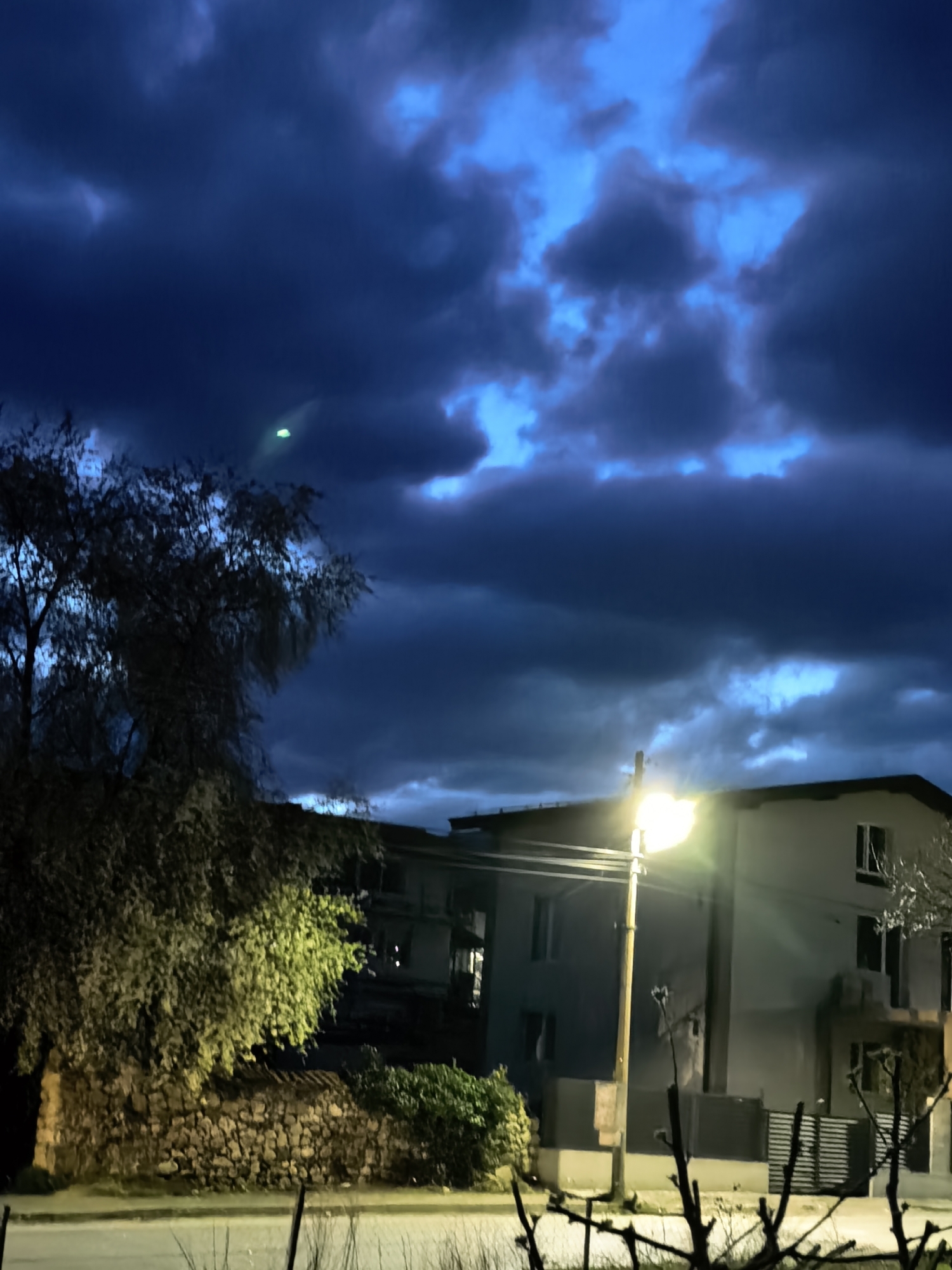The image depicts a street scene at night in the city of Unknown, Unknown. Dominating the view is a dramatic, cloudy sky with shades of deep blue and areas of lighter blue. On the left side, a tree is silhouetted against the sky, and a stone wall is visible beneath the tree. A building is in the background, lit by a bright street lamp in front of it. The street lamp illuminates the building and the surrounding area. In the foreground, there are some leafless branches.