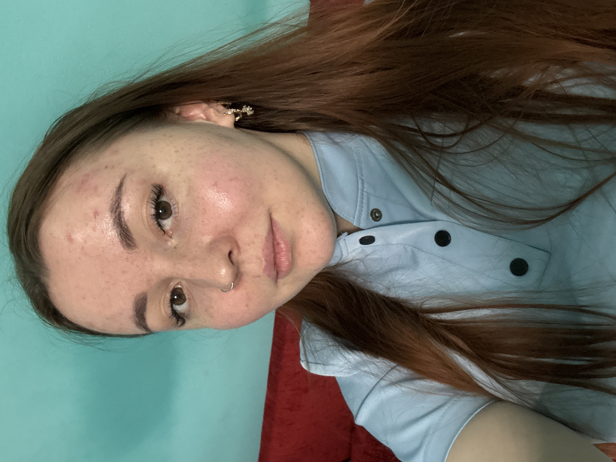 A woman with auburn hair is shown from the chest up, indoors. She wears a light blue, button-down shirt. The background is a combination of teal wall and a red couch or surface. The woman is looking at the camera with a slight smile. She has fair skin with freckles, a nose ring, and earrings. There is no evidence in the image to suggest the location is in Izhevsk, Russia.