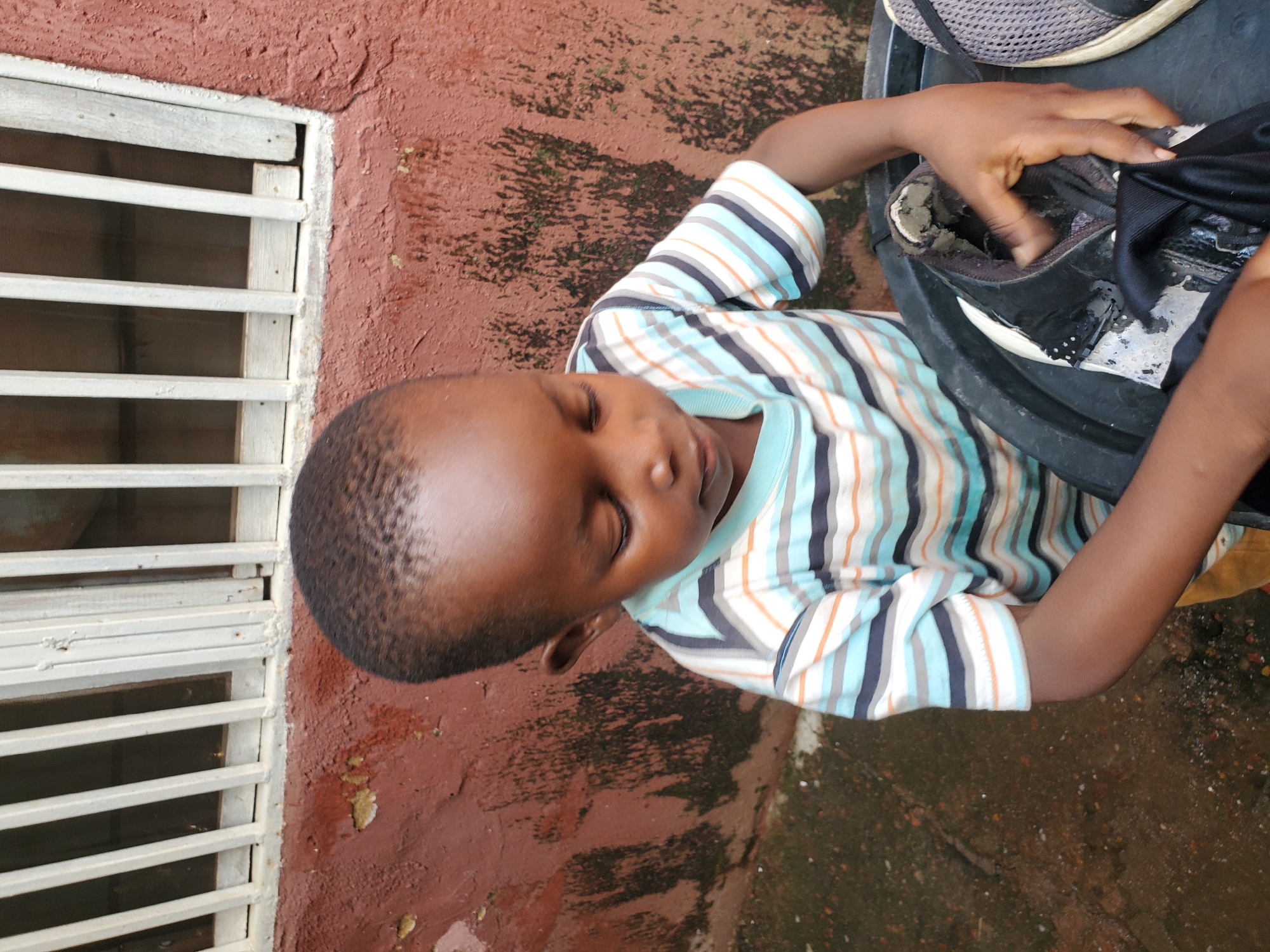 A young boy, with dark skin and short, dark hair, is lying down with his eyes closed. He is wearing a striped t-shirt with blue, orange, white, and dark grey stripes. His right arm is bent at the elbow, and his hand is reaching into a black shoe with a white sole and a grey mesh top. The shoe appears to be worn and dirty. The boy is positioned on a reddish-brown, textured surface that looks like a wall or pavement, with some dark splotches that could be wet patches or moss.
To his left, there is a white louvered window or vent, with horizontal slats. The wall surface is rough and uneven. The lighting suggests it might be daytime, and the overall scene appears to be outdoors, possibly in a residential area. The boy's closed eyes and relaxed posture might indicate he is resting or napping. The location context of Jos, Nigeria, suggests a warm climate. There is no visible text in the image.