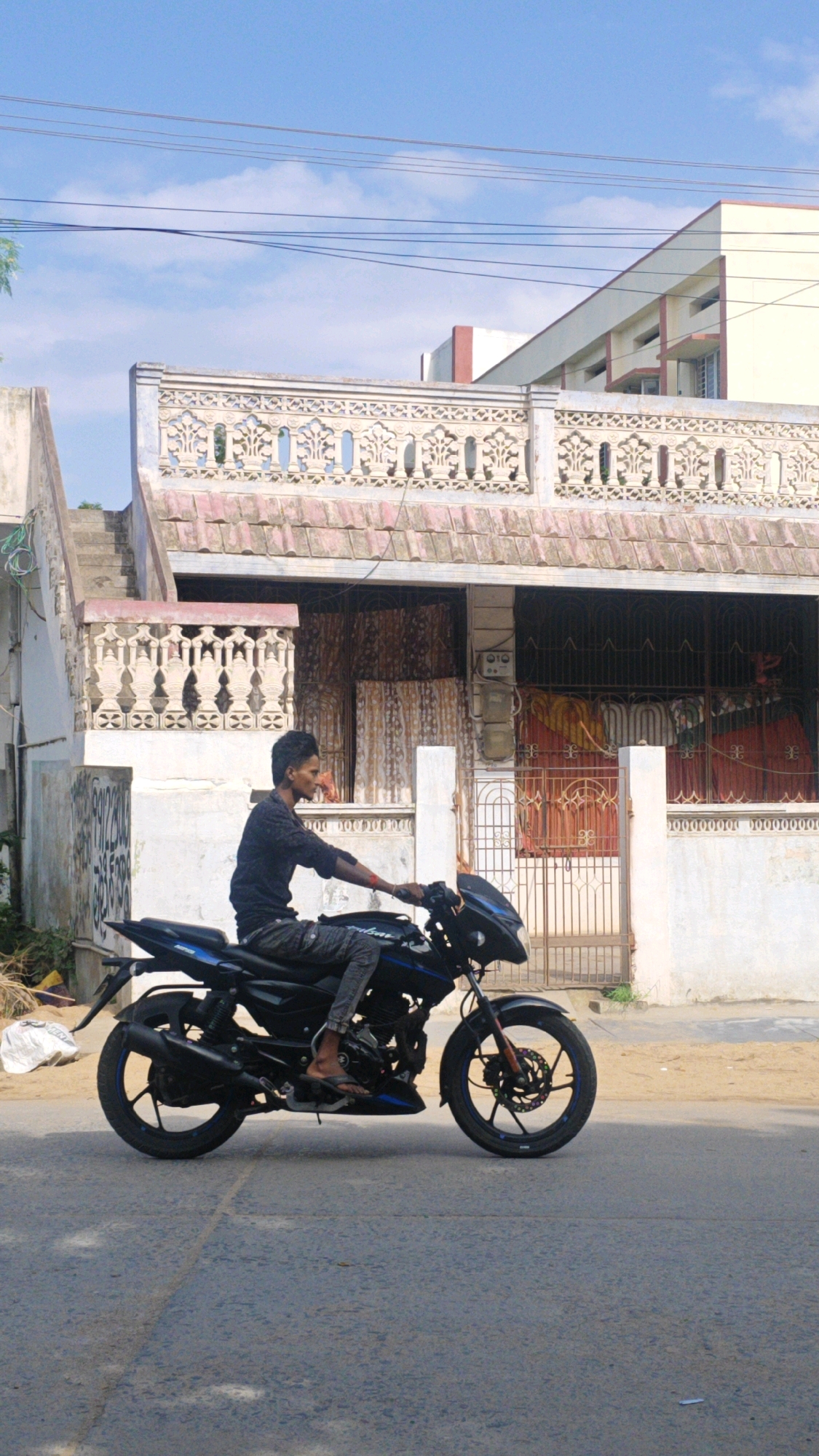 A young man is riding a black motorcycle on a paved street in Pamur, India. The man is wearing a dark long-sleeved shirt and grey pants. He is looking to his right. The motorcycle is in motion, with the front wheel slightly turned. Behind the motorcycle, there is a building with a decorative white railing on its roof. The building has multiple windows and a metal gate in front. The sky is blue with some clouds. The overall environment suggests a residential area in a town.