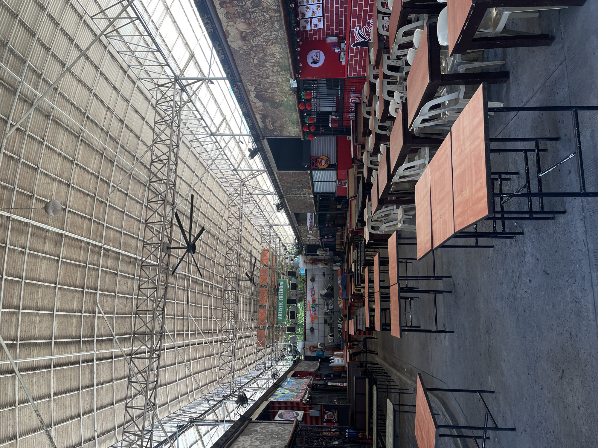 This image depicts an outdoor dining area in Davao City, Philippines, likely a food court or a market with multiple food stalls. The scene is dominated by rows of empty wooden tables with black metal frames and plastic chairs, suggesting it is either early in the day or during a slow period. Above, a large, corrugated metal roof supported by a complex metal framework provides shelter. Several industrial-sized ceiling fans are visible, indicating a warm climate.
Along the sides of the open-air space, various food stalls are set up. One stall features a stage with musical instruments and signage that reads "ARTISTIC FREEDOM." Another stall is adorned with red brick-patterned siding and displays a menu board. Large murals depicting historical or cultural scenes are painted on the walls of some of the stalls, adding a touch of local artistry. Red paper lanterns hang above some of the stalls, suggesting a festive atmosphere or an upcoming event. The ground is paved with concrete. No people are actively engaged in dining or any other activity within the frame. The overall impression is of a large, open-air venue prepared for customers but currently unoccupied.