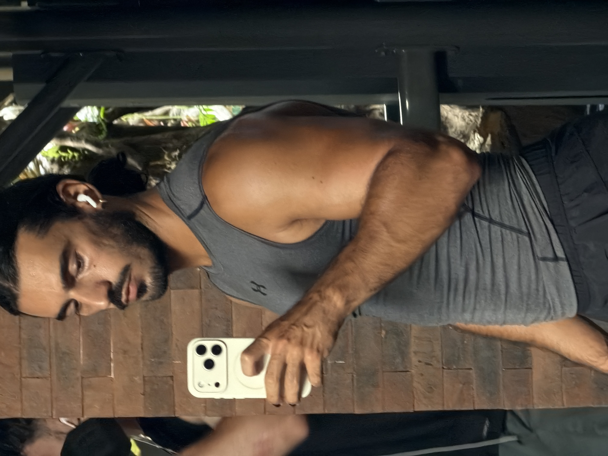 A man with dark hair pulled back, a mustache, and a beard takes a mirror selfie in what appears to be an outdoor gym or fitness area. He is wearing a grey sleeveless Under Armour shirt and black shorts. He holds a white smartphone in his left hand, which is pointed towards the mirror. In his right ear, he wears a white wireless earbud. The background shows a brick wall, some foliage, and metal gym equipment. The scene is well-lit, possibly in the daytime. The location is identified as North Kuta, Indonesia.