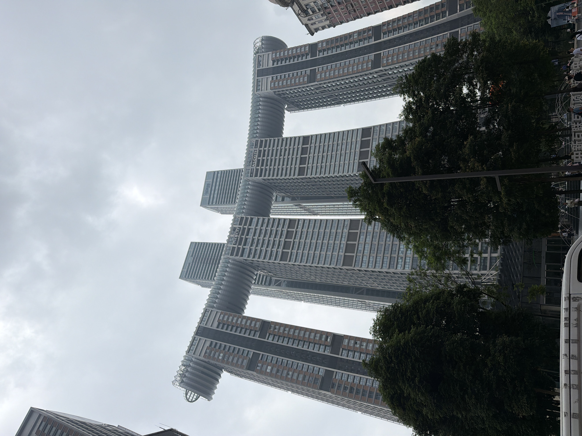 The image shows a low-angle shot of the Raffles City Chongqing, a mixed-use skyscraper complex located in the Yuzhong District of Chongqing, China. The complex features eight skyscrapers, with four of them connected by a horizontal structure known as "The Crystal" or "The Ribbon." This horizontal element bridges the four towers and houses amenities such as observation decks, restaurants, and a sky garden. The architecture is modern and striking, with the towers varying in height and design.
The sky is overcast and grey, suggesting a cloudy day. In the foreground, lush green trees are visible, partially obscuring the lower levels of the buildings. A street lamp stands on the right side of the image, near the trees. In the very bottom of the frame, the top portion of a white van is visible, along with a street sign and what appears to be part of a pedestrian walkway with some people walking. The overall impression is one of a modern, urban landscape with significant architectural innovation.