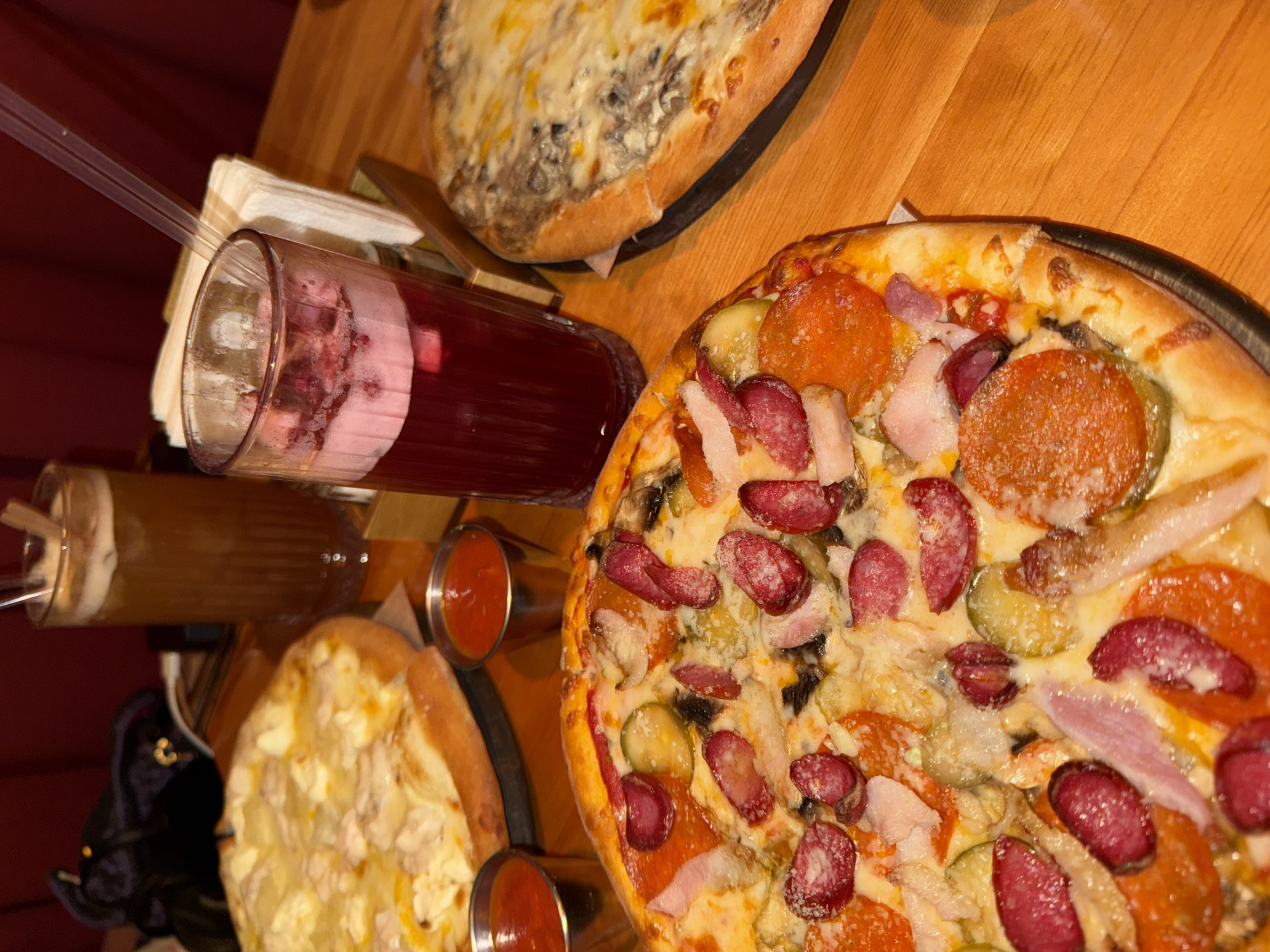 This image captures an overhead, angled view of a dining table laden with food and drinks, likely in a restaurant or cafe in Perm, Russia. The scene is illuminated by warm, artificial lighting.
The primary subject is a large, round pizza, occupying the right side of the frame. This pizza features a golden-brown crust and a generous covering of melted cheese, pepperoni slices, small, dark red salami or sausage pieces, light-colored ham or bacon bits, and distinct green slices of pickled cucumbers. Underneath the toppings, dark specks suggest the presence of mushrooms.
To the bottom left, partially visible, is another pizza with a creamy white topping and chunks, possibly chicken or mushrooms. Further in the background, another pizza with a similar light-colored topping is seen.
Accompanying the food are two tall glasses with beverages. One glass, prominently placed near the top left, contains a dark red drink with a frothy, pinkish layer on top and what appear to be crushed berries, served with a clear straw. Another glass, partially obscured on the left, holds an amber-colored liquid, also with a straw.
Small metal ramekins filled with a red sauce, likely ketchup or marinara, are scattered on the table. A stack of white napkins rests near the red drink. The table itself is made of light-colored wood with visible grain. A dark red background, possibly a wall or curtain, frames the left side of the scene. A black object, possibly a bag or coat, is partially visible at the bottom left. No people are visible, and there is no discernible text. The arrangement suggests a casual and inviting meal.