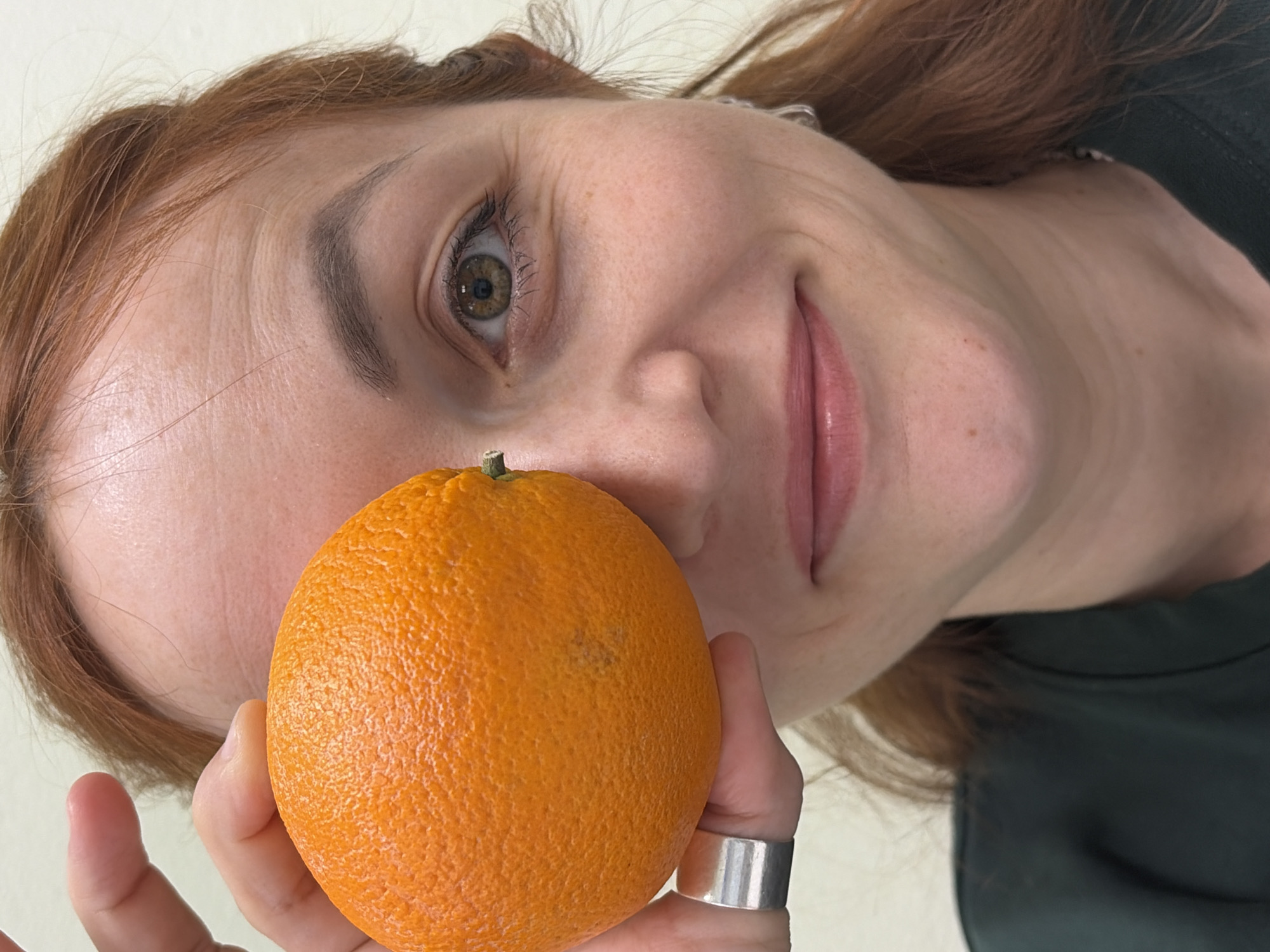 A woman with light skin and red hair is holding an orange in her hand, positioning it near her face. She is wearing a silver ring on her finger. The background is a plain, light color, possibly a wall or ceiling. The image perspective is tilted. The specific location in the city of Chaikovsky, Russia, is not discernible from the visual information provided.