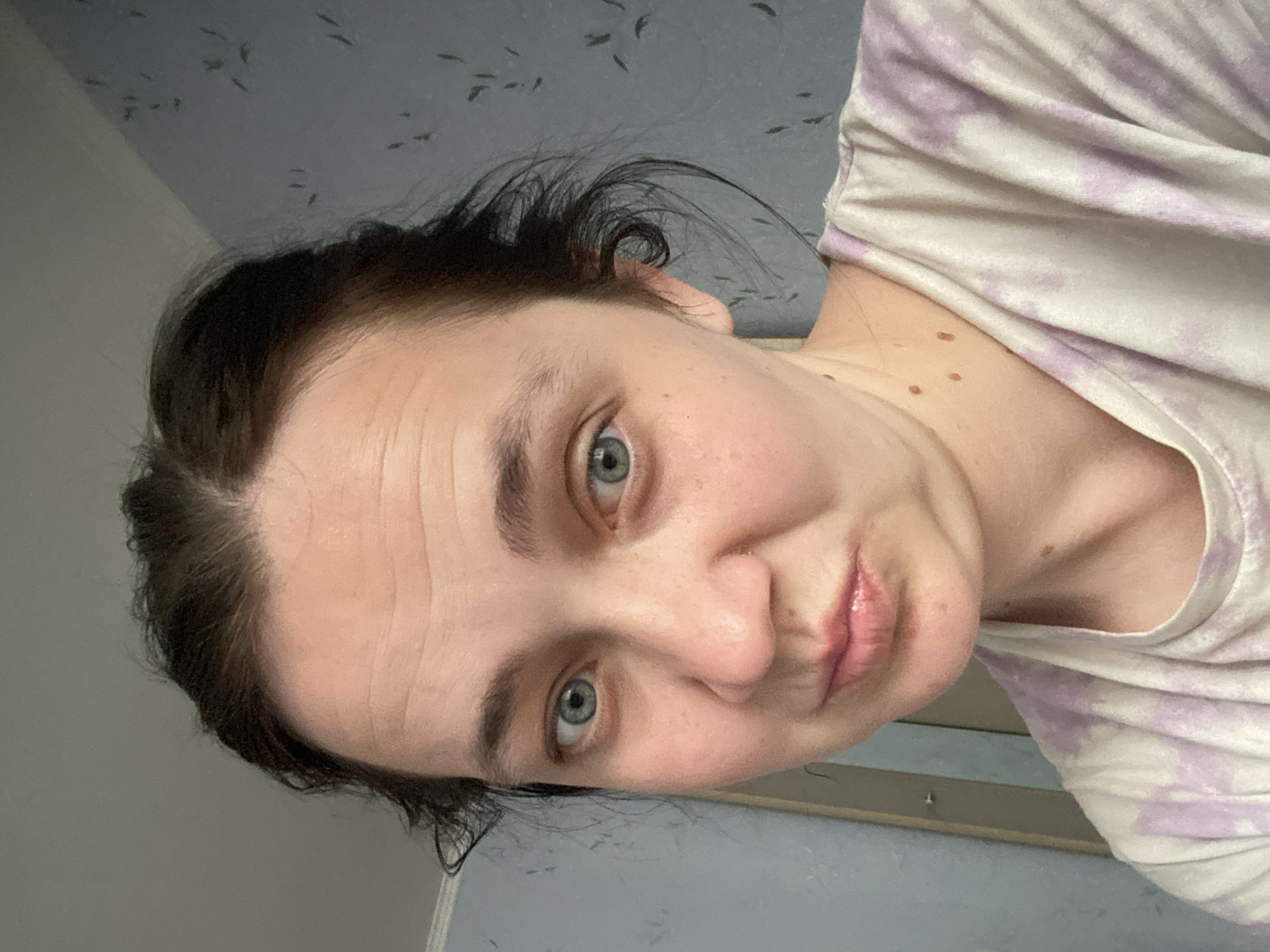 A woman is pictured from the shoulders up, looking directly at the camera with a slight pout. She has dark brown hair pulled back, revealing a forehead with visible wrinkles. Her eyes are a light blue-green, and her skin is fair with some freckles and moles visible on her neck. She is wearing a t-shirt with a tie-dye pattern in shades of white and light purple. The background is a blue wall with a subtle floral or patterned design. The lighting appears to be artificial, coming from the front and slightly to the left.