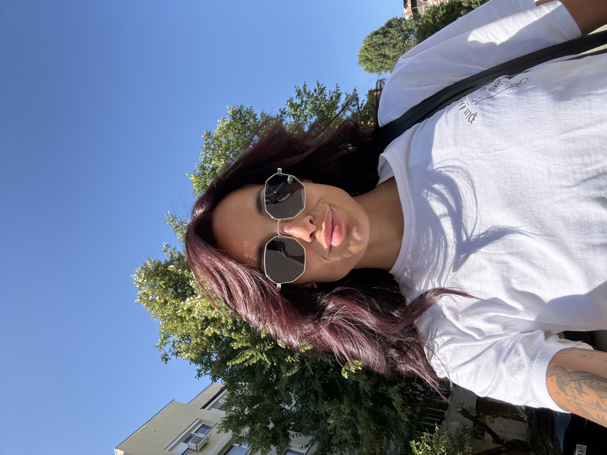A young woman with burgundy hair and sunglasses is taking a selfie outdoors. She is wearing a white t-shirt. Behind her, a building and trees are visible under a clear blue sky. The location is the city of Pleven, Bulgaria.