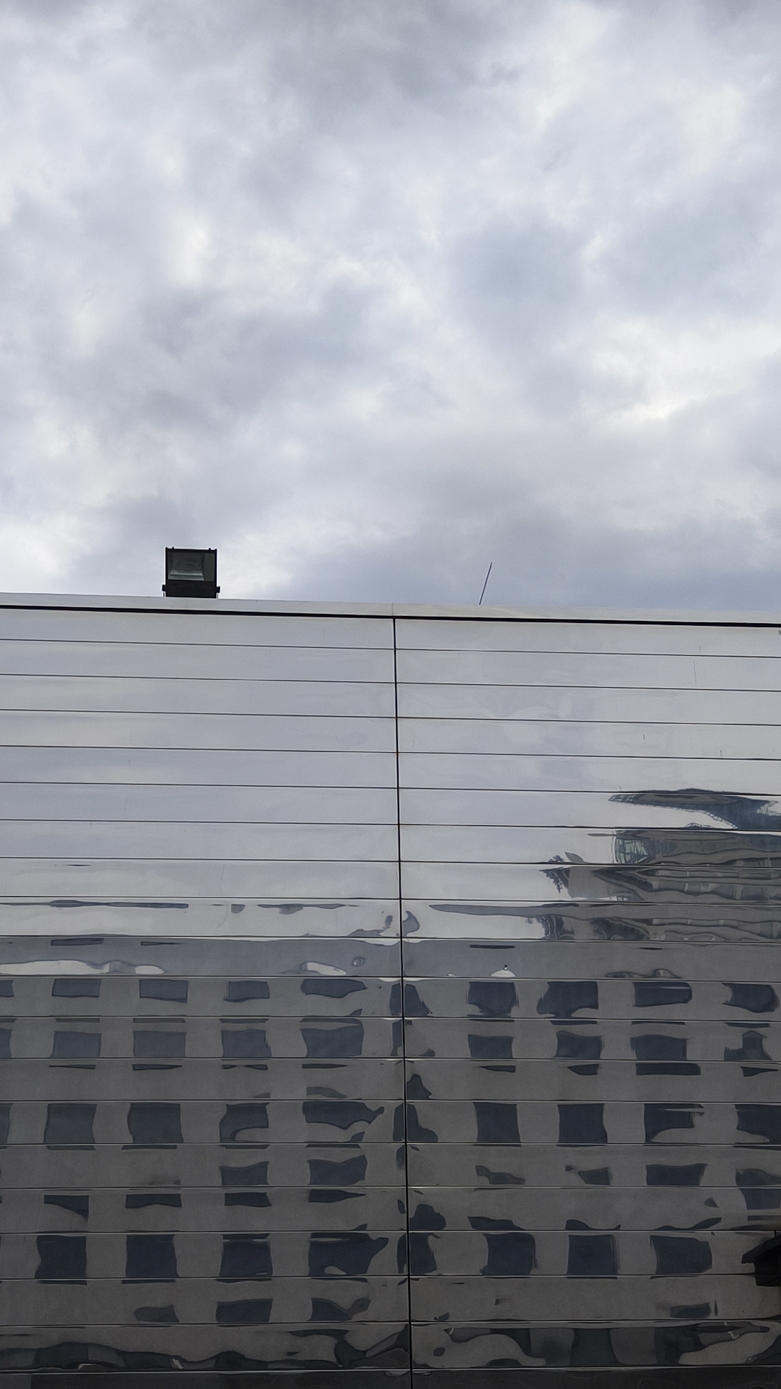 This is a vertical, eye-level, medium shot of a modern building's facade under a cloudy sky. The building is covered in horizontal, reflective metal panels. The upper part of the facade reflects the overcast sky. The lower part of the facade reflects a distorted image of a building with a grid pattern of windows. On the rooftop, there is a rectangular spotlight with a dark housing. A thin, vertical antenna or rod juts out from the rooftop on the right side of the frame. The building appears to be part of a contemporary architectural design, possibly in an urban setting. The scene evokes a sense of muted light and geometric reflection.