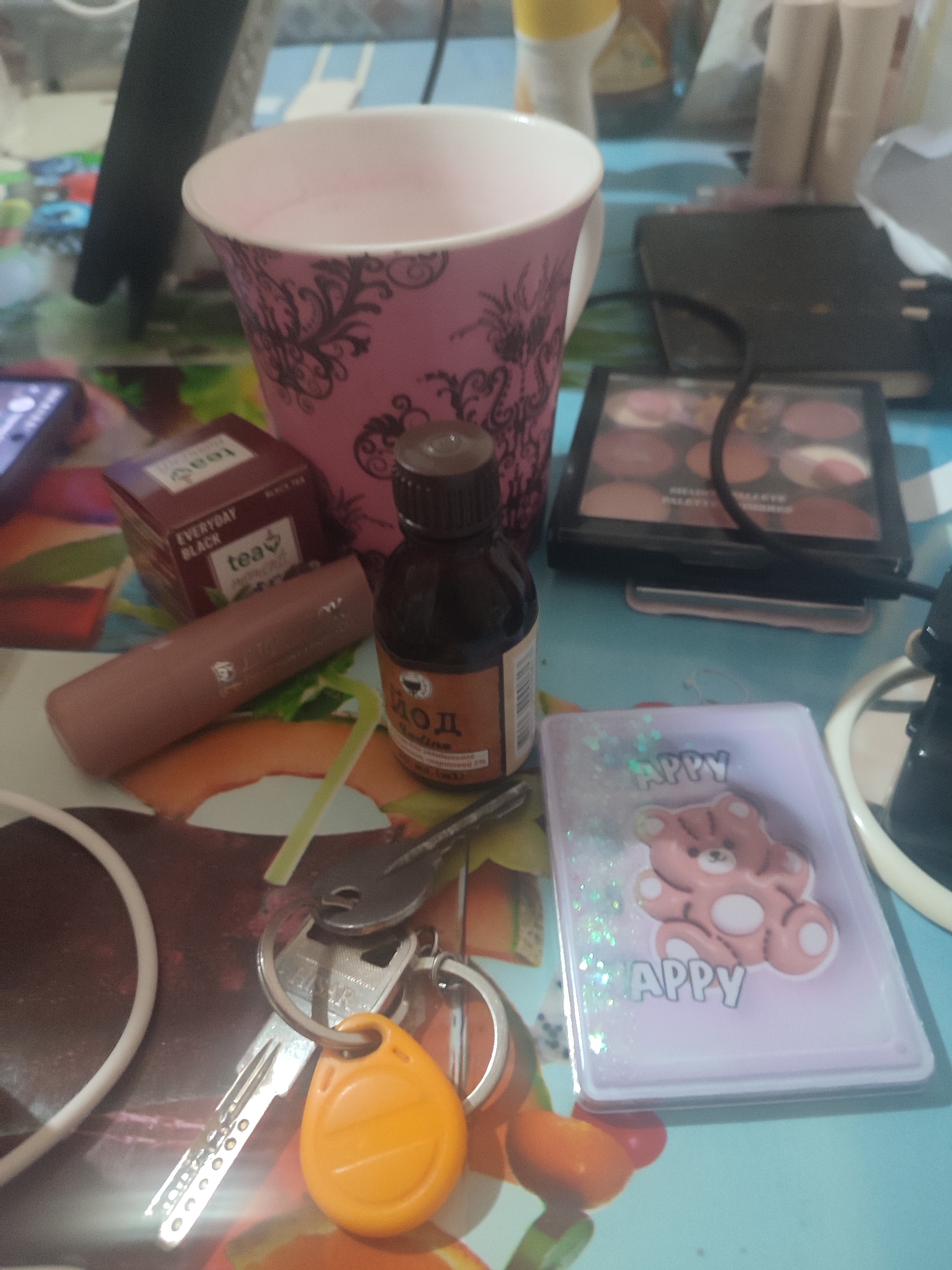 The image shows a flat lay arrangement of various personal items on a patterned surface, likely a table. In the center is a pink decorative mug with black filigree, partially filled with a light pink liquid. To its right is a small brown glass bottle with a dark cap, labeled in Cyrillic script with "йод" (iodine). Next to the bottle is a lipstick or balm in a muted pink casing. Behind these items, a black eyeshadow palette with multiple circular pans is visible. In the lower left quadrant, a set of keys with an orange fob is arranged on a white circular cord. To the right of the keys, a small translucent purple case with a glittery interior and a pink teddy bear sticker adorned with the word "HAPPY" is positioned. The overall scene suggests a personal vanity or desk area. The image does not contain any explicit information about the city of Chernivtsi, Ukraine, or any specific location within it.
