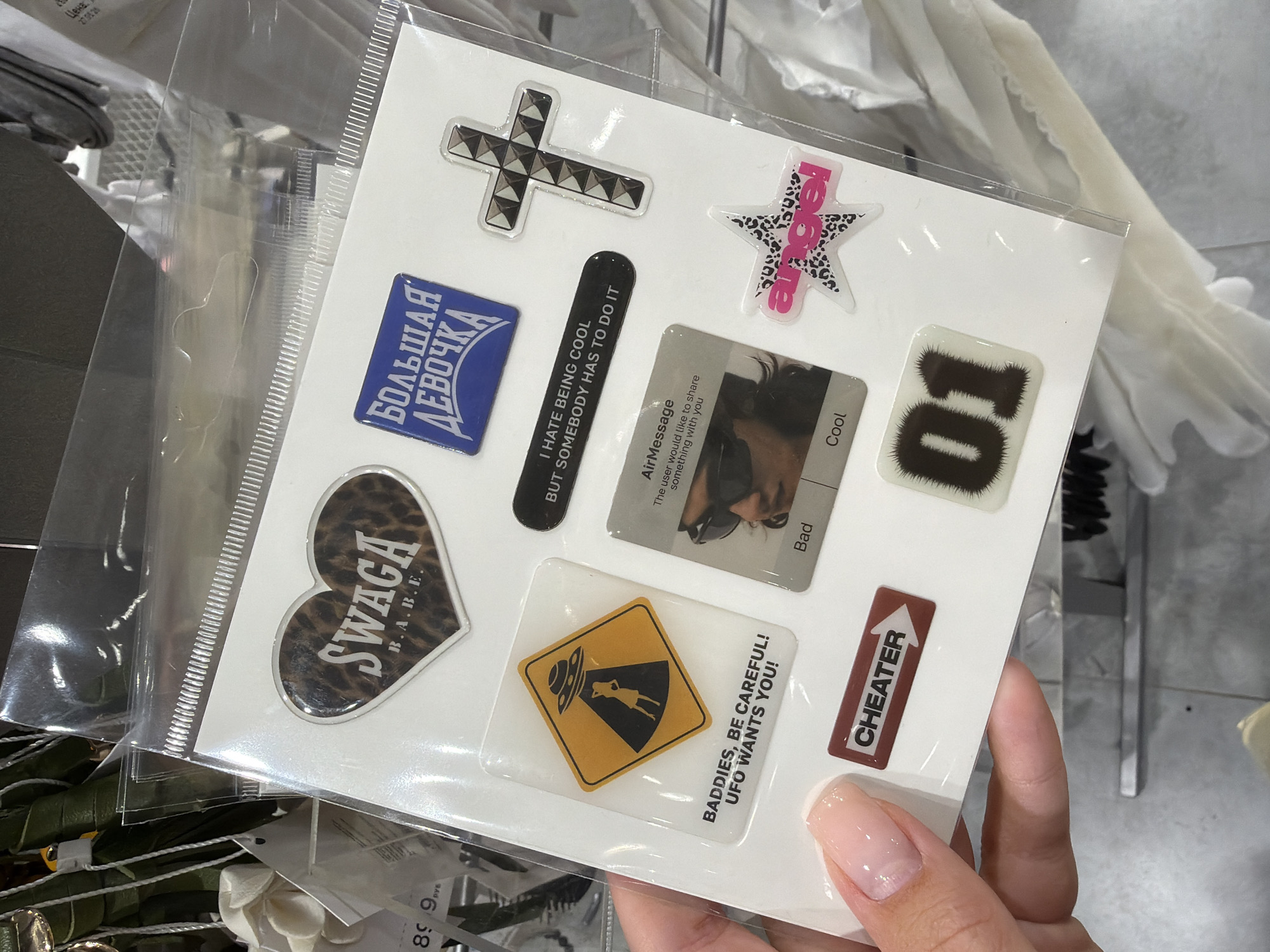 This media file displays a sheet of stickers being held by a person's hand. The stickers are of various shapes and designs, including a cross, a star with "angel" text, a blue rectangle with Russian text that translates to "BIG GIRL," a black oval with English text that reads "I HATE BEING COOL BUT SOMEBODY HAS TO DO IT," a heart with leopard print and "SWAGA B.A.B.E." text, a yellow diamond warning sign with an alien and a figure being abducted, a rectangle with "CHEATER" text and an arrow, a rectangular sticker with a person's face and the text "AirMessage The user would like to share something with you Bad Cool," and a square sticker with the number "01" and a furry texture. The background shows clothing items on racks, suggesting the stickers are being displayed in a retail setting, possibly a clothing store in Moscow, Russia. The lighting appears to be indoor lighting. The hand holding the stickers has a light pink nail polish. Visible price tags in Russian currency (руб) are also present in the background.