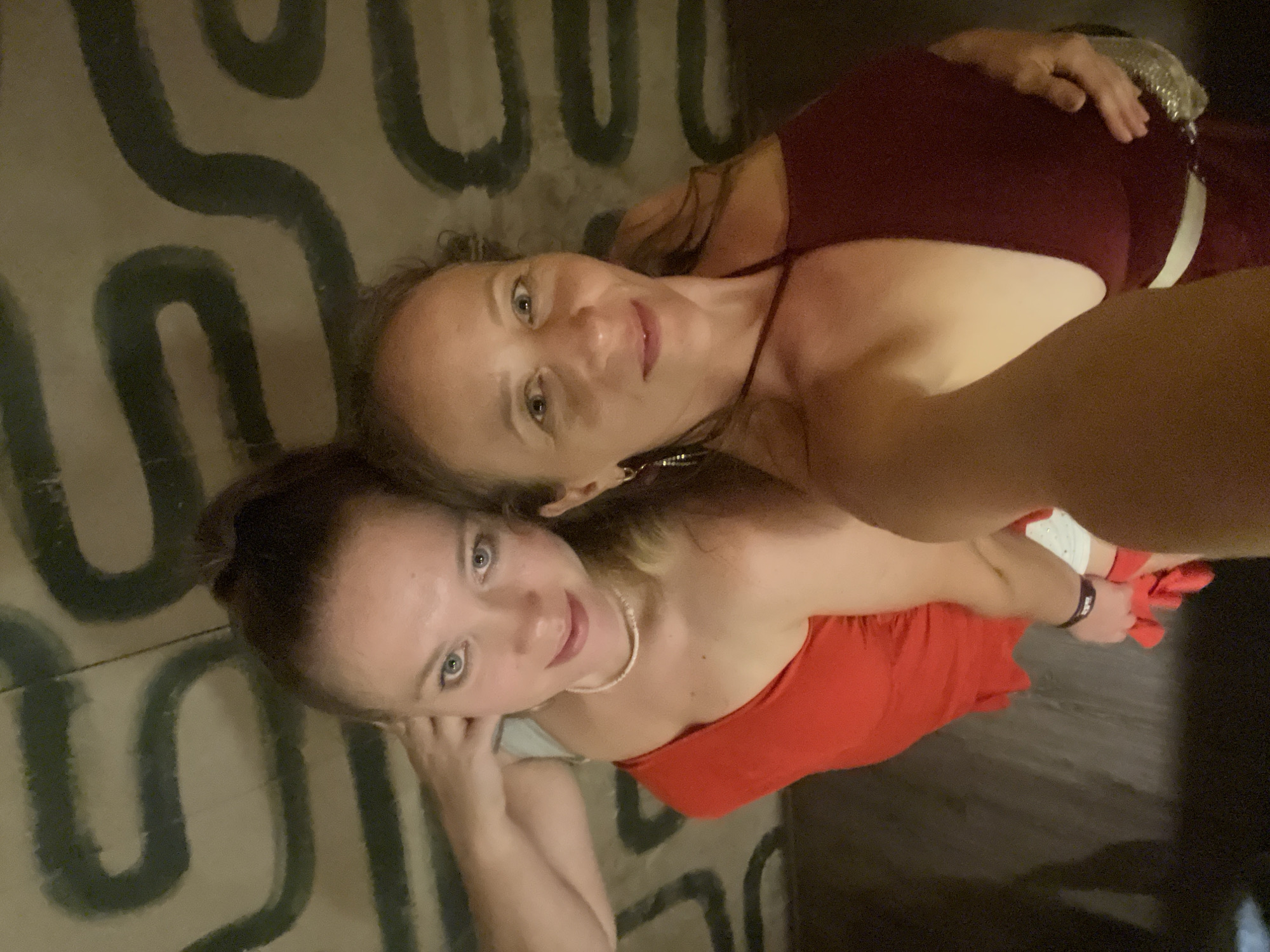 Two women are taking a selfie indoors. The woman on the left is wearing a red off-the-shoulder dress and a pearl necklace. Her hair is pulled up in a ponytail. The woman on the right is wearing a dark red halter top. The background appears to be a wall with a patterned design. The floor is dark and textured. No specific location in Đà Nẵng, Vietnam can be identified.