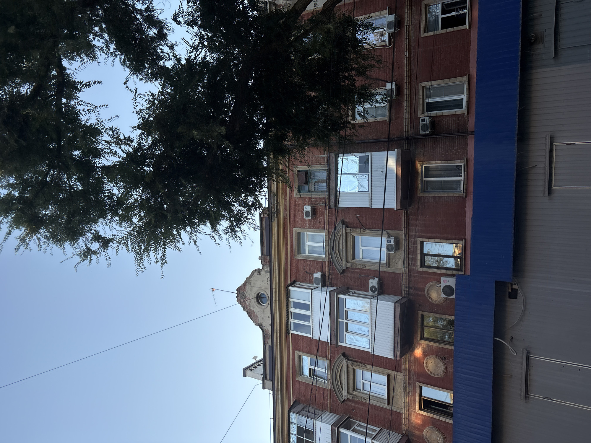 This is a low-angle exterior, medium shot of a brick apartment building with a blue awning in the foreground, taken in Rostov-on-Don, Russia. The building has multiple floors with windows and balconies. Some windows are open and some are closed. There are air conditioning units visible on the building's facade. A large tree with dark green foliage partially obscures the right side of the building. Overhead, a clear blue sky is visible with a single power line stretching across it. The top of the building features a damaged pediment with a circular window. The overall impression is one of an urban residential area.