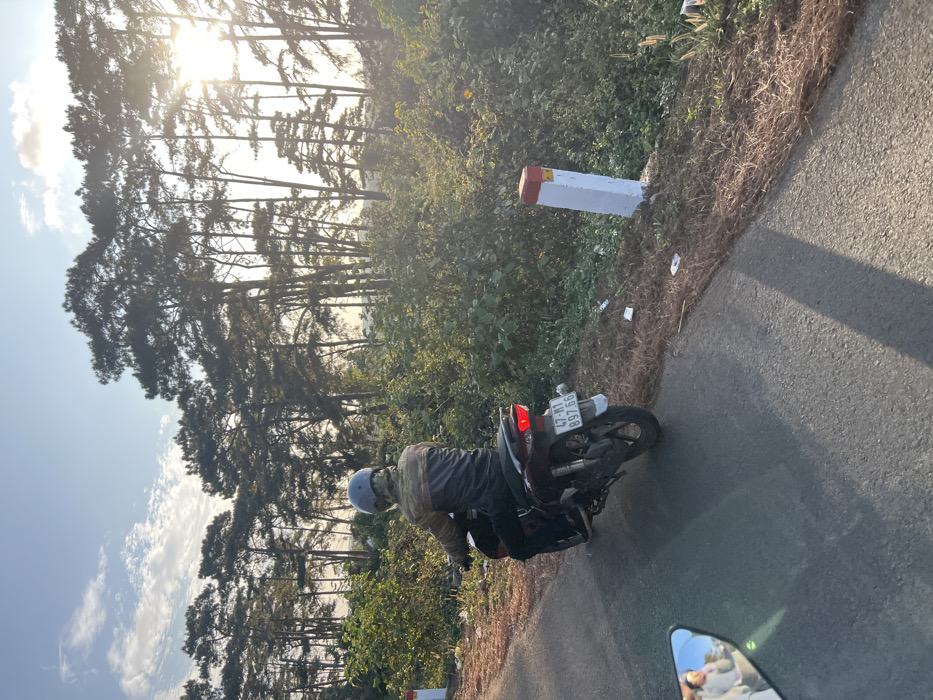 An outdoor scene in Ea Ksô, Vietnam, depicts a rural asphalt road under late afternoon or early morning sunlight. A solitary individual, wearing a gray helmet and a camouflage-patterned jacket, operates a dark scooter/motorcycle. The motorcycle, displaying Vietnamese license plate "47-N1 897.66" and an illuminated red taillight, travels away from the camera perspective, positioned on the left side of the road. Long shadows from unseen objects stretch across the road from left to right. The roadside features dense green foliage, dry brown grass, and scattered white litter. A white concrete marker post with a red top is visible on the right side of the road. A line of tall, slender trees, consistent with pine trees, forms the distant horizon. The sun, partially obscured by trees, emits a strong glare from the upper right, casting a warm light over the scene. A portion of a motorcycle rearview mirror is visible in the lower-left foreground, reflecting blurred figures.