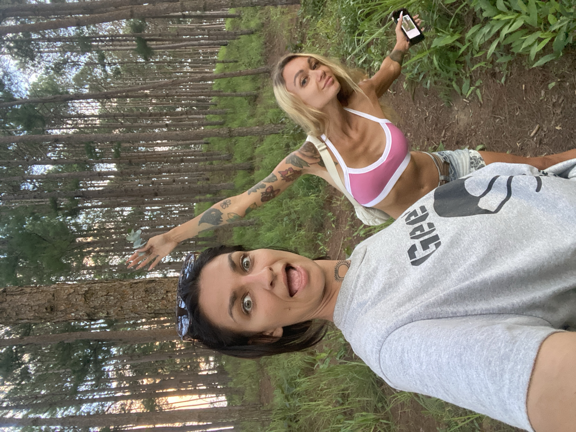 In an outdoor scene with tall, slender trees forming a dense backdrop, two individuals are captured. The image is oriented vertically, suggesting a selfie-style photograph.
In the foreground, a person with dark hair and wide, expressive eyes is looking directly at the camera, their mouth open in a playful or surprised expression. They are wearing a grey t-shirt with a graphic design on the chest, and sunglasses are perched on their head. A tattoo is visible on their neck.
Behind them, another person with blonde hair and numerous tattoos on their arm is leaning back, extending their arms. They are wearing a pink and white sports bra and denim shorts. In their right hand, they hold a smartphone, seemingly capturing the moment. A light-colored strap from a bag is visible over their shoulder.
The setting appears to be a forest or wooded area, with a path visible between the trees. Lush green grass and foliage line the path. The lighting suggests it might be daytime, possibly late afternoon given the quality of light filtering through the trees. The overall mood of the image seems lighthearted and candid, with both subjects displaying engaged and energetic emotions.