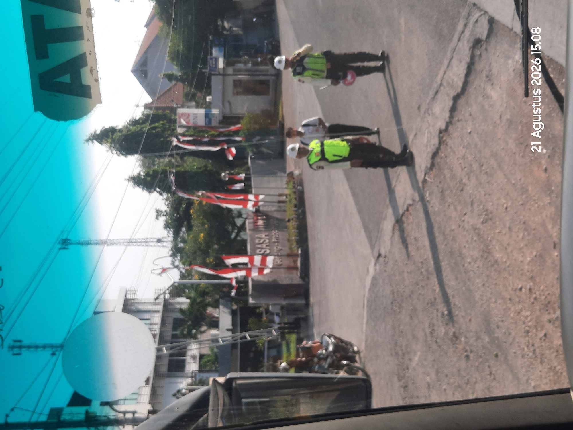 The image shows a street scene in Gending, Indonesia, with two security guards in high-visibility vests and hard hats directing traffic. One guard is holding a red traffic cone. A third person, possibly a civilian, stands behind them. In the background, there is a sign that reads "SASA PINTU" and Indonesian flags are displayed. A motorcycle is parked to the left of the frame. The sky is a bright cyan color, suggesting it may be a reflection or tinted window. The timestamp "21 Agustus 2026 15.08" is visible in the lower right corner.