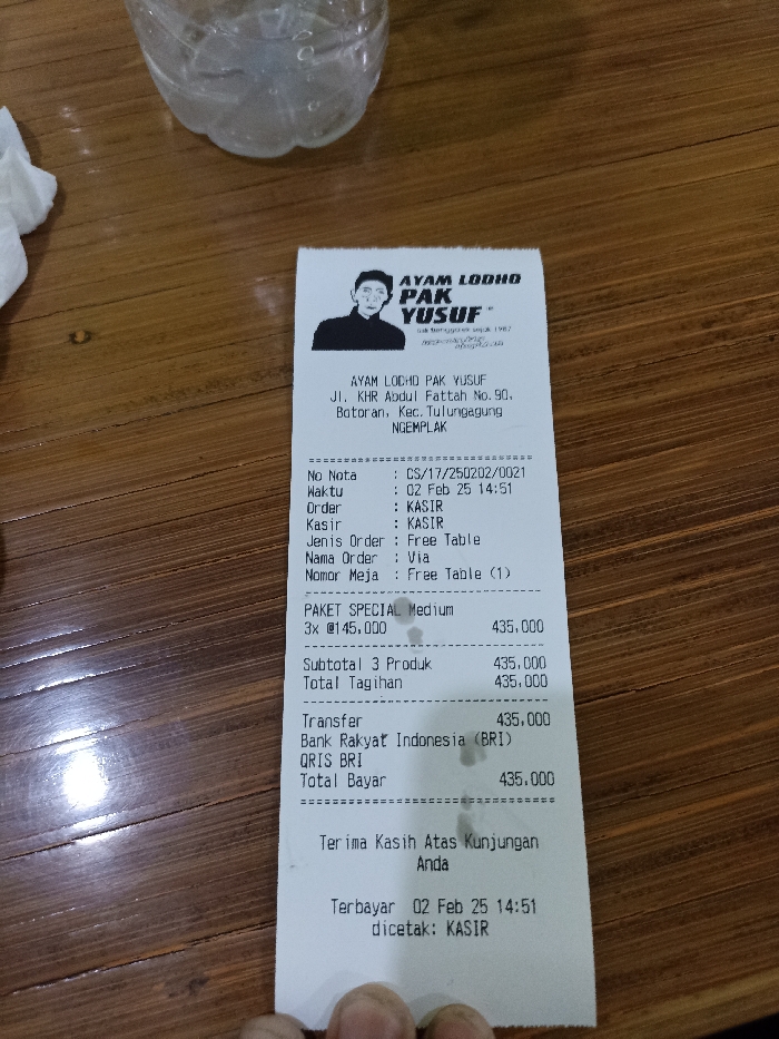 A close-up, high-angle shot captures a printed receipt resting on a wooden table, likely in a restaurant setting. Part of a clear plastic water bottle and a white crumpled napkin are visible in the background on the left.
The receipt, from "AYAM LODHO PAK YUSUF," displays its address as "Jl. KHR Abdul Fattah No. 90, Botoran, Kec. Tulungagung NGEMPLAK," situating the location in Tulungagung, Indonesia. The logo at the top features a stylized drawing of a man's head and shoulders.
The transaction details indicate a note number CS/17/250202/0021, and the time of purchase and printing as 02 Feb 25 14:51 (February 25th, 2:51 PM). The order type is "Free Table," and the cashier is "KASIR."
The purchased item listed is "PAKET SPECIAL Medium," with 3 units priced at @145,000 each, totaling 435,000. The subtotal for "3 Produk" (3 Products) and the "Total Tagihan" (Total Bill) are both 435,000. The payment was made via "Transfer" through "Bank Rakyat Indonesia (BRI)" and "QRIS BRI," with the "Total Bayar" (Total Payment) also recorded as 435,000.
The receipt concludes with a "Terima Kasih Atas Kunjungan Anda" (Thank You For Your Visit) message, confirming the payment time and that it was "dicetak: KASIR" (printed by: CASHIER).