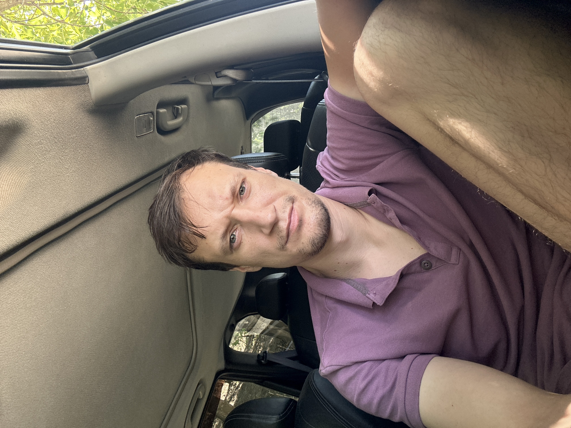 A man is sitting in the driver's seat of a car, his body angled towards the camera. He is wearing a purple polo shirt. The car's interior is visible, including the headliner, sunroof, and black leather seats. Sunlight streams in through the windows, highlighting his leg. The scene appears to be outdoors, with trees visible through the car windows. There is no indication of the location being Makiv, Ukraine.