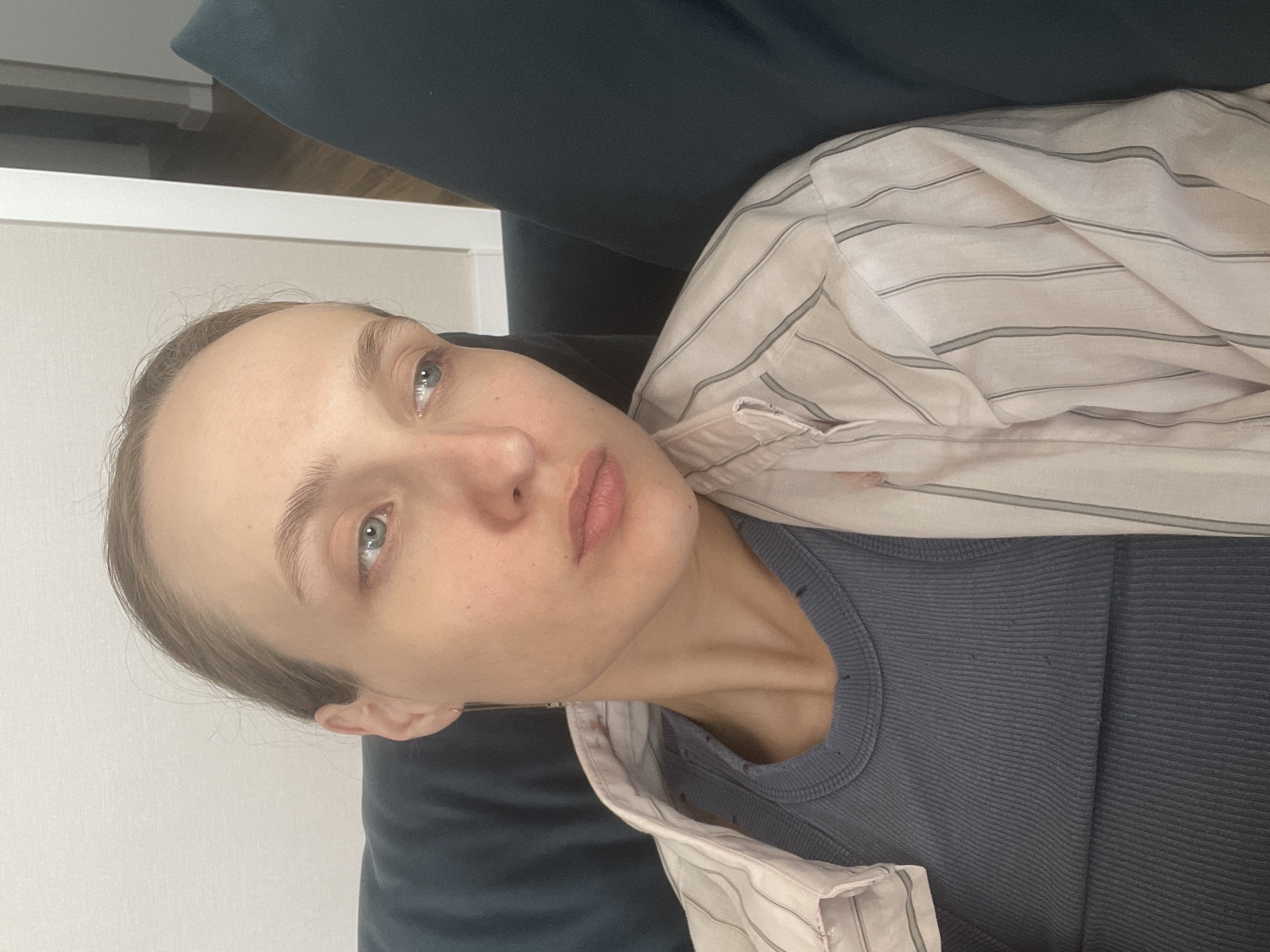 A pale-skinned individual with light brown hair and blue eyes lies in an indeterminate location, possibly indoors. The person wears a dark grey ribbed shirt and a light-colored, striped button-down shirt. The individual is partially obscured by what appears to be a dark blue sofa. The background consists of a white wall. The image is oriented sideways. There is no indication of the location being Sepych, Russia.