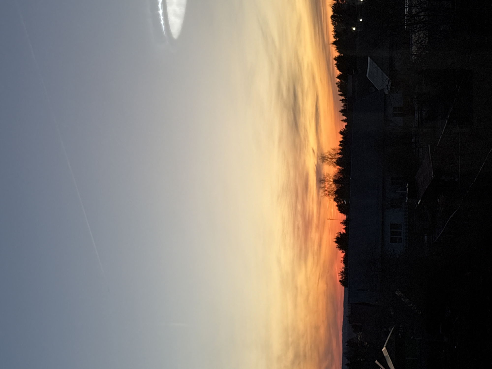 The image shows a sunset over what appears to be a residential area. The sky transitions from blue to orange and yellow near the horizon. There are buildings and trees silhouetted against the bright sky. The location is likely Прикамский, Russia, based on the image context.