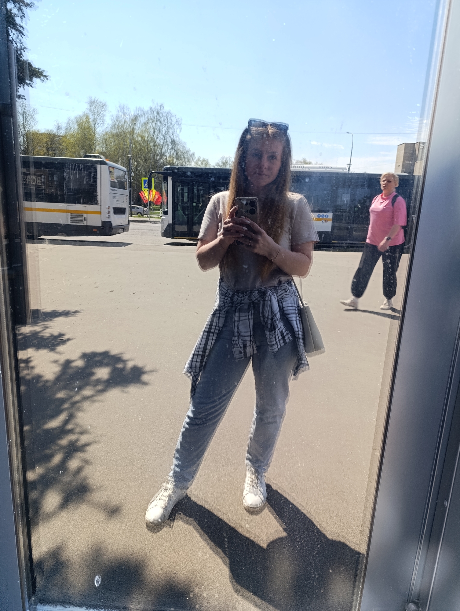 A young woman is taking a selfie in a mirror. She is wearing a light-colored t-shirt, light wash jeans, white sneakers, and a plaid shirt tied around her waist. She is also wearing sunglasses on her head and carrying a light-colored purse over her shoulder. In the reflection, a bus station is visible with two buses and a woman in a pink shirt walking. The scene is outdoors on a sunny day.