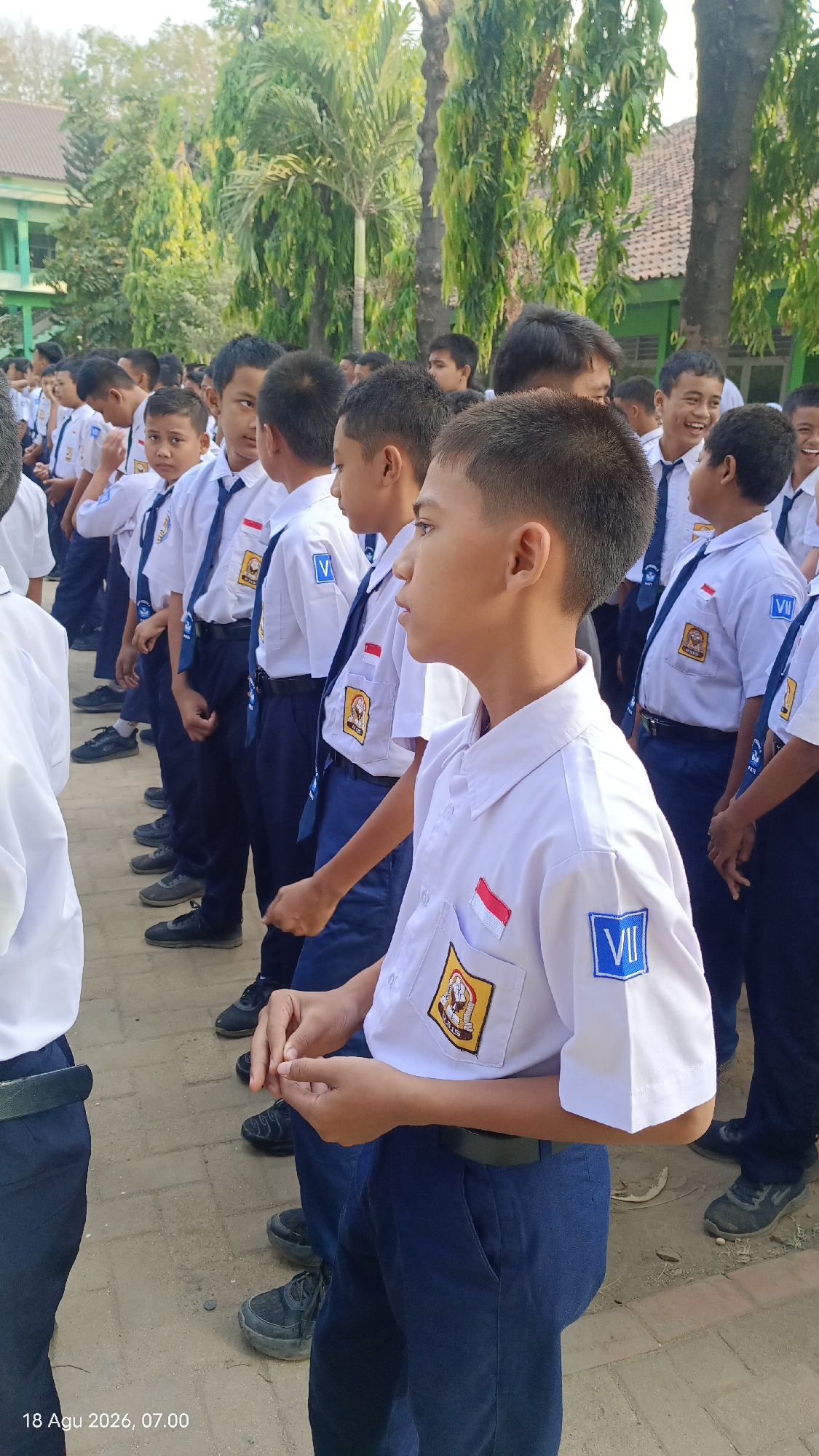 In Kecamatan Pati, Indonesia, a group of male students in white school uniforms with blue ties stand in formation outdoors. The students are positioned in multiple rows, facing to the right of the frame. They are wearing dark blue pants and black shoes. On their left sleeves, some students have patches with a "VII" in a blue square, and on their chests, they have school emblems. The background features trees, buildings, and a tiled roof. The ground is paved with concrete and dirt. The timestamp "18 Agu 2026, 07.00" is visible in the bottom left corner.
