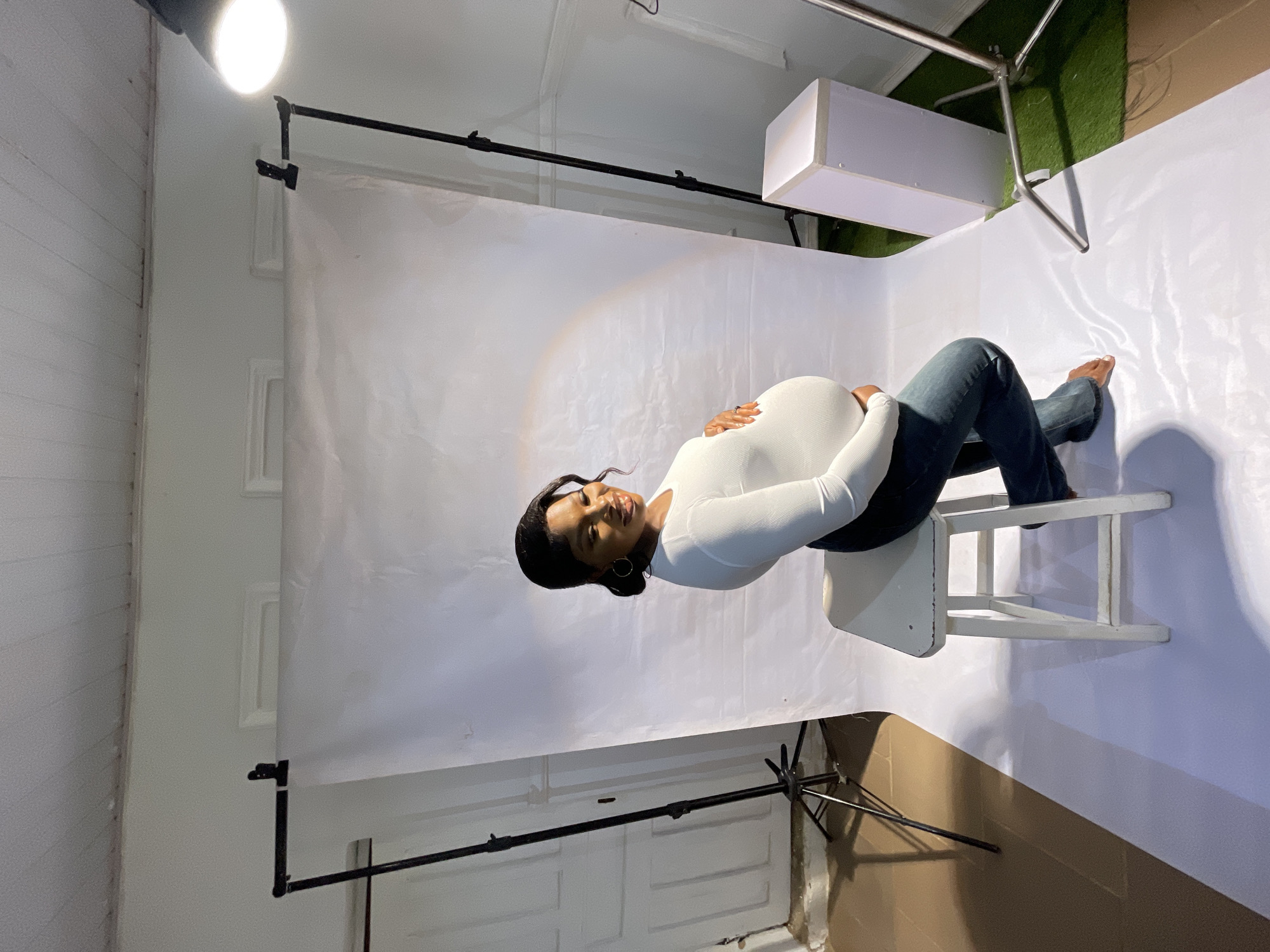 A woman is lying on her left side on a white wooden chair, facing the camera. She is positioned in a studio setting with a white backdrop and lighting equipment. She wears a long-sleeved white shirt and dark blue jeans. The studio setting appears to be indoors, with white walls and flooring. Additional photography equipment is visible, including light stands and a large white light box. The location cannot be precisely determined as Jos, Nigeria, from visual information alone.