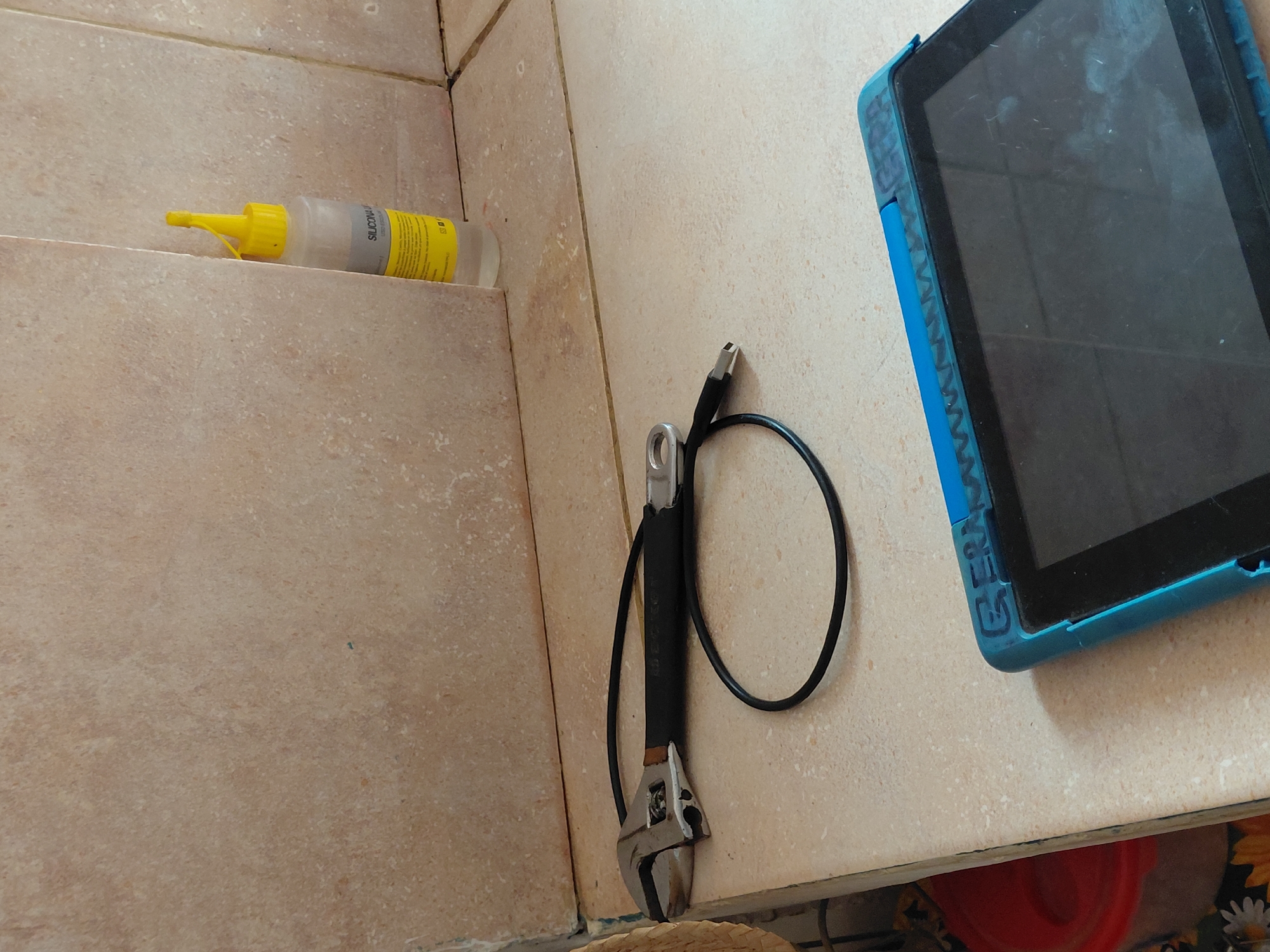 This media file captures an indoor scene featuring various objects arranged on and beside light brown, speckled tiled surfaces. The composition shows a lower tiled ledge and a flat tiled surface, possibly a counter or shelf, with a tiled wall in the background.
On the lower tiled ledge to the left, a clear plastic squeeze bottle with a bright yellow nozzle and cap is placed horizontally. A yellow label is wrapped around the bottle, with the word "SILICONA" partially visible, indicating its contents are likely silicone sealant or glue. Additional, unreadable text is also present on the label.
On the main tiled surface, an adjustable wrench with a black rubber handle grip is seen. A black charging cable is looped around the wrench, with one end, a USB connector, clearly visible. To the right, a dark-screened tablet is laid on the surface. It is protected by a textured, bright blue case, on the side of which the partial word "GERMA" is visible, possibly part of a brand name. The tablet screen shows noticeable smudges and fingerprints.
No human presence or specific activity is depicted; the items appear to be stationary. The lighting is even, suggesting an indoor setting. Below the main surface, a hint of a red object and a floral-patterned item (possibly a fabric or basket) can be seen. The general appearance of the items suggests practical use, and the tiles indicate a utilitarian space, such as a bathroom, kitchen, or workshop. The image does not provide specific visual cues to confirm the Santa Ana, Venezuela location beyond being an indoor space.