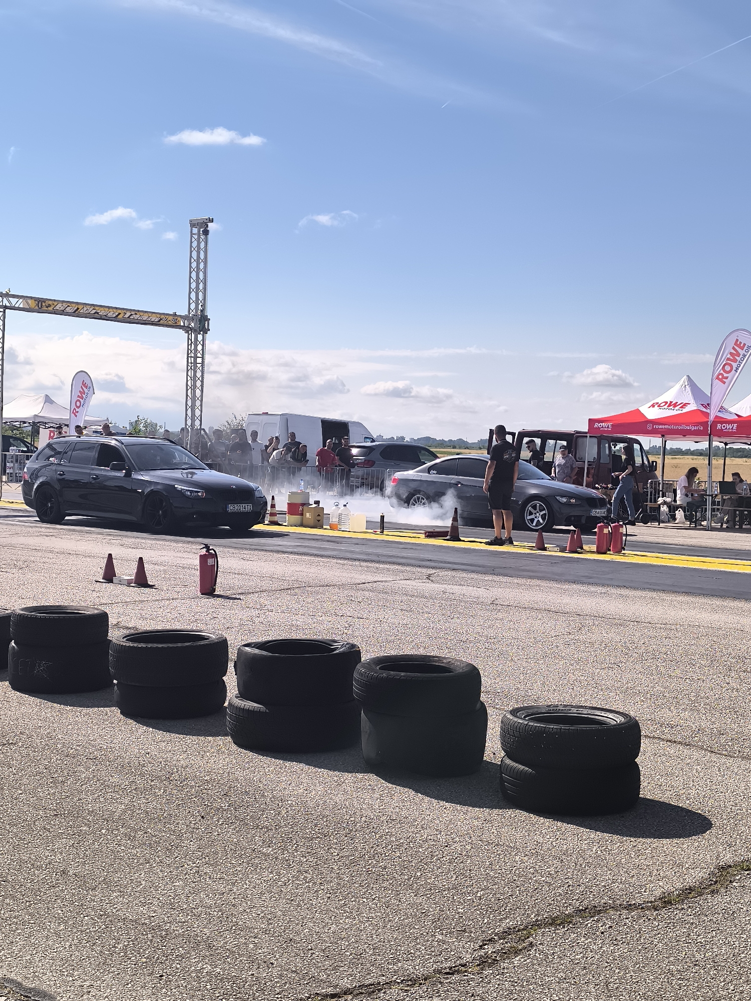At the Akatsievo Drag Racing track in Bulgaria, a drag race is underway. Two cars, a black station wagon and a dark gray coupe, are positioned side-by-side on the asphalt track. Both vehicles are emitting smoke from their tires, indicating they are revving their engines. Several people are standing around, observing the race. In the background, there are tents and a white van. The sky is clear and blue with a few scattered clouds. Piled tires are visible in the foreground, likely used as a safety barrier.
