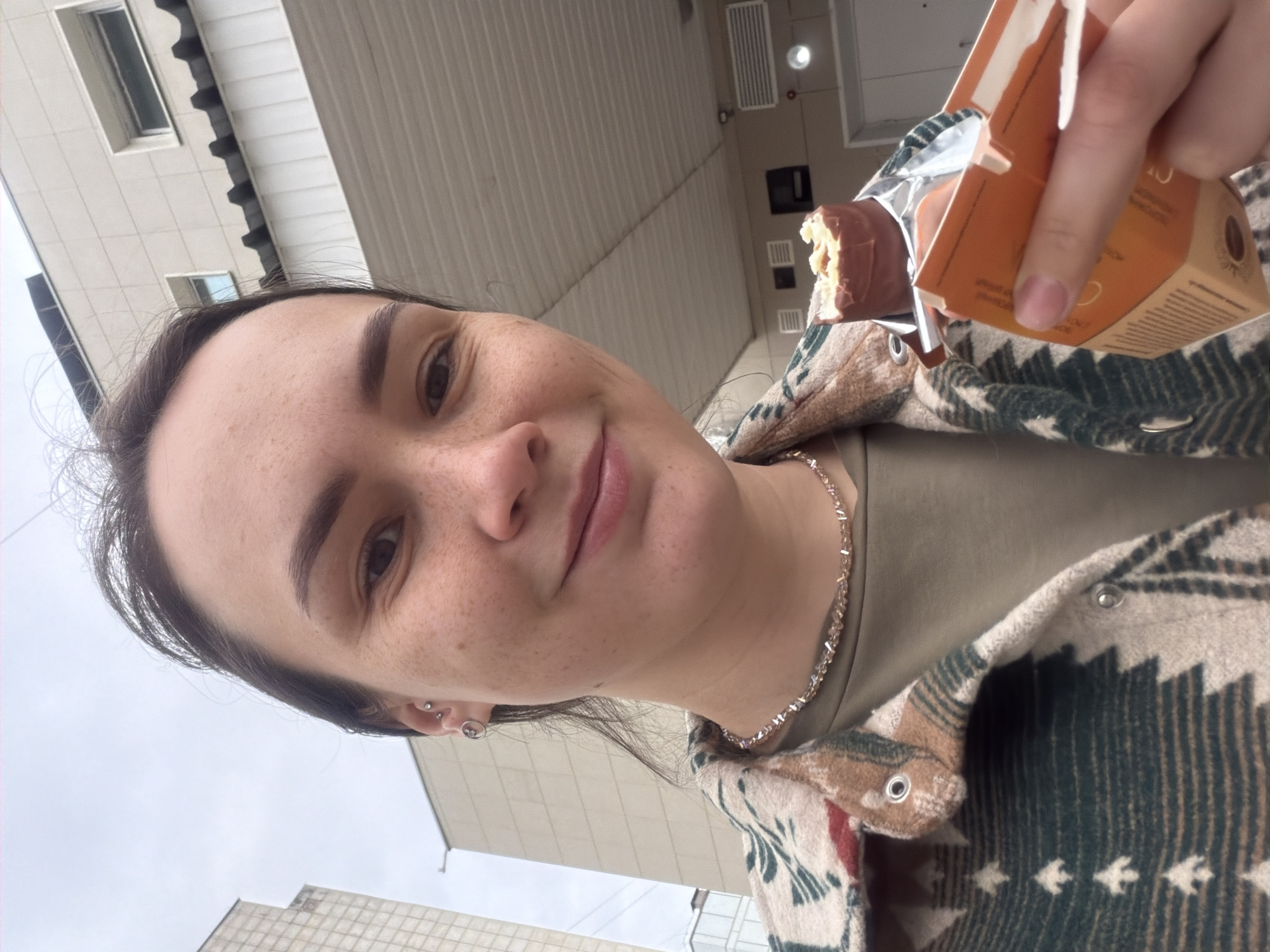 A medium shot shows a light-skinned woman with dark hair and freckles holding an unwrapped candy bar next to its opened package. The woman is wearing a dark green t-shirt and a patterned overshirt with buttons. She also has a silver necklace and earrings.
The background includes a building with white siding and windows. The image was likely taken in an urban area, potentially in the city of Chaikovsky, Russia. The sky is overcast.